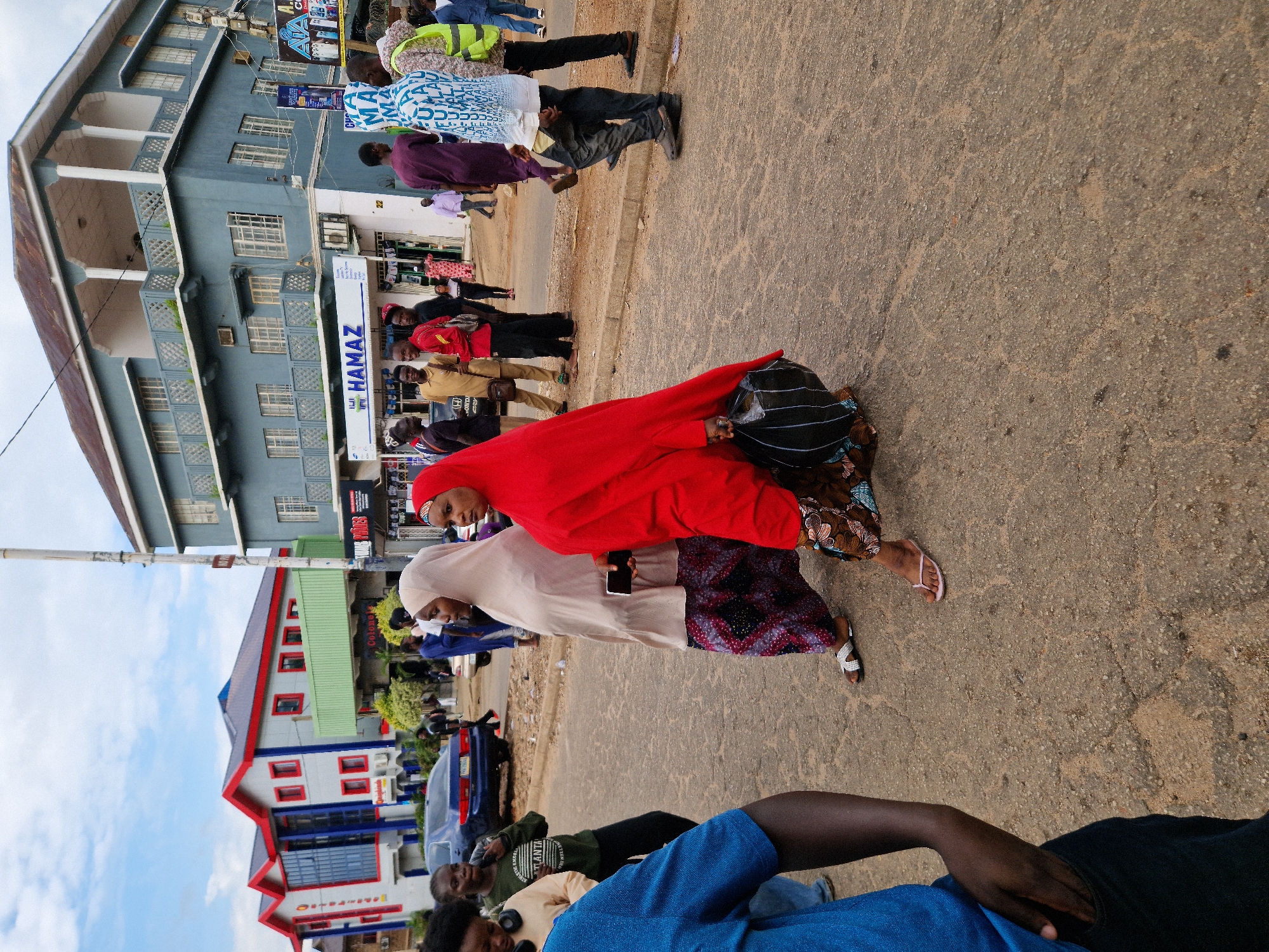 The image depicts an outdoor scene on a paved street, likely in a commercial area, under an overcast sky. Several individuals are present, some walking and others standing. Two women in the foreground are prominently featured, one wearing a beige hijab and a patterned skirt, and the other in a vibrant red hijab and a patterned skirt, carrying a black bag. To their left, a person in a blue shirt is partially visible, with their arm in their pocket. In the background, a blue car is parked on the street. A multi-story building with light blue walls and balconies stands behind the people, with a business called "HAMAZ" visible on the ground floor. Another building with a red and gray facade and red trim is visible further to the left, with a green awning extending from it. The ground is a rough, gray asphalt surface with some visible cracks and patches. The overall impression is a snapshot of daily life in a potentially urban environment.