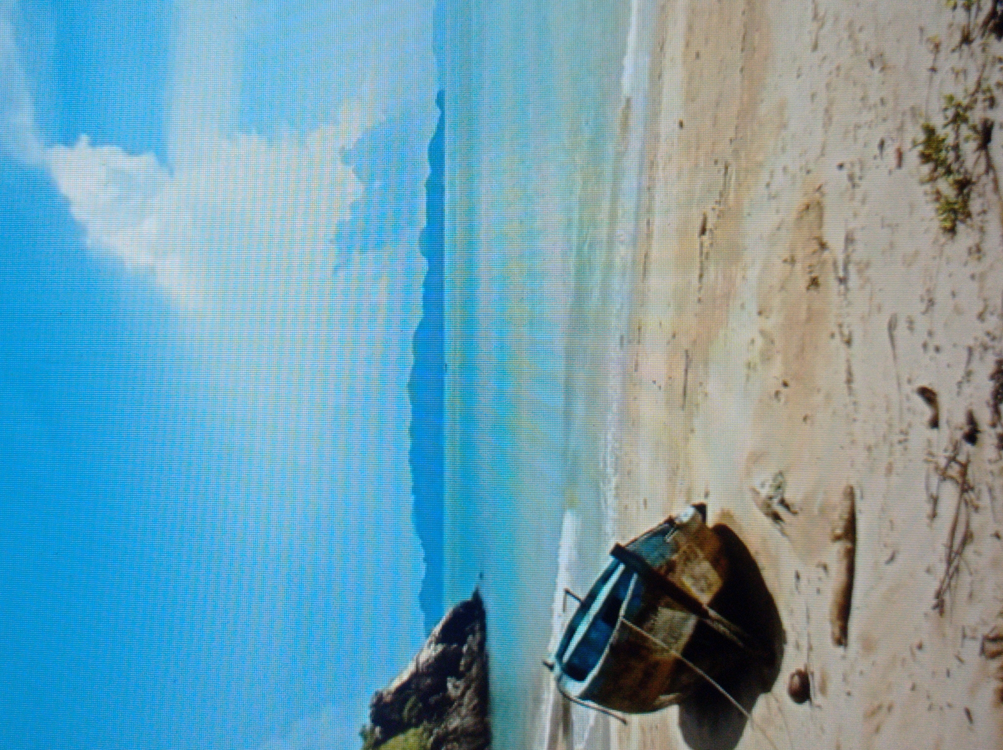 This is a photograph of a tranquil beach scene. The image shows a sandy beach with a small, weathered boat resting on its side near the water's edge. The water is a clear, bright blue, with gentle waves lapping at the shore. In the distance, a chain of dark, low-lying islands or mountains can be seen against the horizon. The sky is a vibrant blue, with a few scattered white clouds, one of which is particularly prominent and fluffy. The scene evokes a sense of peace and isolation.
The question mentions the city of Mukachevo, Ukraine. However, this image depicts a coastal scene with islands and a body of water that appears to be an ocean or large sea, which is not characteristic of Mukachevo, a landlocked city in Ukraine. Therefore, the location is not Mukachevo, Ukraine.