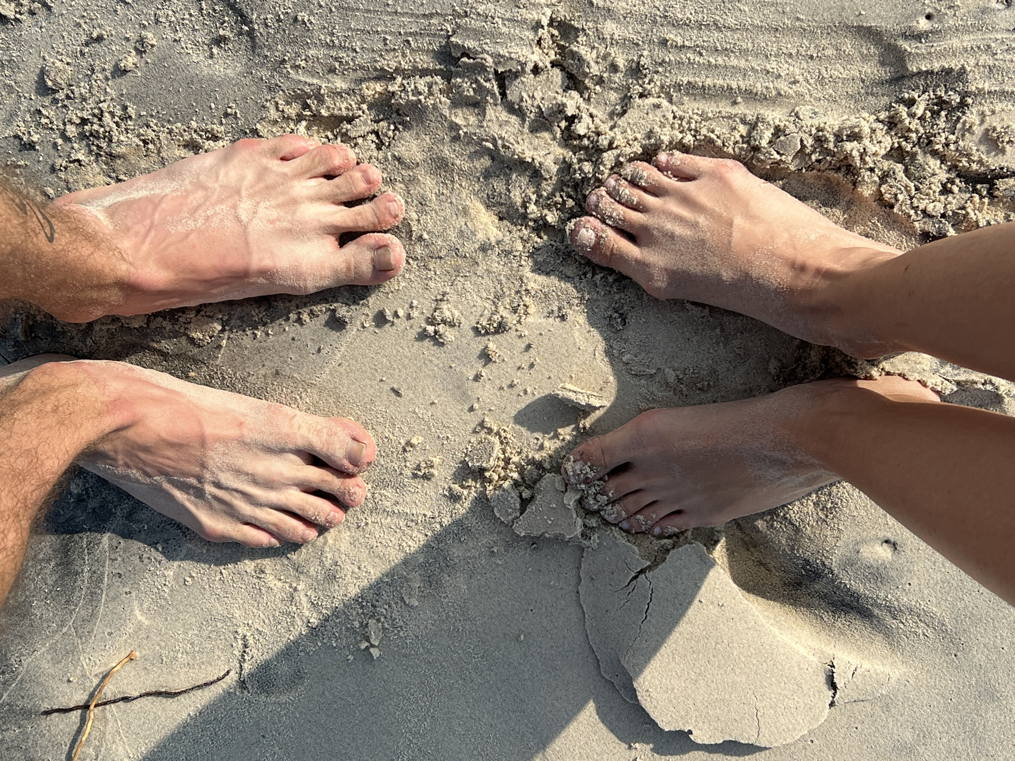 An overhead shot captures two pairs of feet resting on the sandy ground, likely on a beach in Nam Nha Trang, Vietnam. The scene is predominantly light-colored sand, showcasing varied textures from fine grains to slightly coarser, clumpy sections, and some areas where the sand appears damp and compacted.
On the left side of the frame, a pair of male feet and lower legs are visible, characterized by the presence of hair and prominent veins. These feet are partially coated with dry sand, and a faint tattoo or mark is visible on the ankle. To the right, a pair of female feet are seen, appearing smoother, smaller, and with less pronounced veins, also lightly dusted with sand. Both sets of feet create subtle indentations in the soft surface.
The strong, distinct shadows cast by the feet and variations in the sand's texture indicate bright, direct sunlight, suggesting it is likely mid-day or early afternoon. The overall ambiance conveyed is one of relaxation and connection, with the individuals presumably enjoying the warmth and natural environment of the beach. A small, dark, thin piece of organic debris, resembling a twig or root, is visible in the lower-left corner of the frame. There is no visible text in the image.