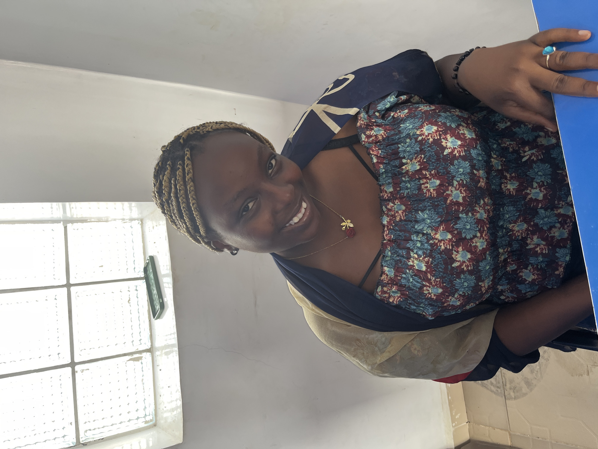 The photograph shows a woman inside a room with white walls and a white window with a clear glass block insert. The woman has dark skin, blonde braids, and a gold necklace, and is wearing a floral patterned shirt and a dark shawl. She is smiling at the camera. The edge of a blue table is visible on the right side. The location is Gura Top, Nigeria.