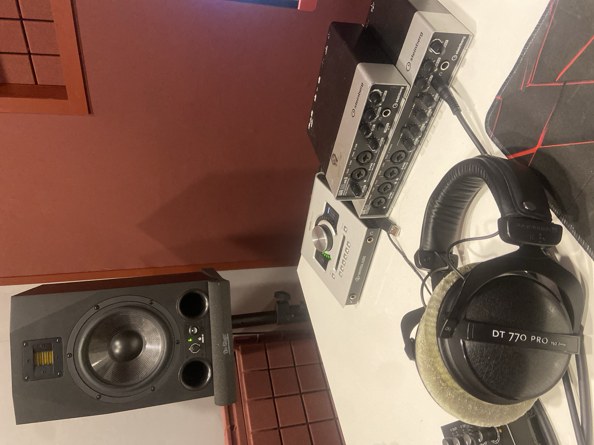 This is a medium shot, eye-level, indoor photograph of a desk in a recording studio. The studio appears to be located in Kazan, Russia, as indicated by the presence of "Kazan" written on a piece of equipment. The background consists of a reddish-brown wall and acoustic foam panels.
On the left side of the image is a black studio monitor speaker on a stand. In the center of the image, on the desk, are several pieces of audio equipment. From left to right, there is a silver Universal Audio Apollo Twin audio interface, followed by two stacked silver Steinberg UR22mkII audio interfaces. A pair of black Beyerdynamic DT 770 PRO headphones are placed in the foreground, partially obscuring the audio interfaces. A black mousepad with red accents is visible on the right side of the desk. Cables are connected to some of the equipment. The overall impression is one of a functional and organized audio recording workspace.