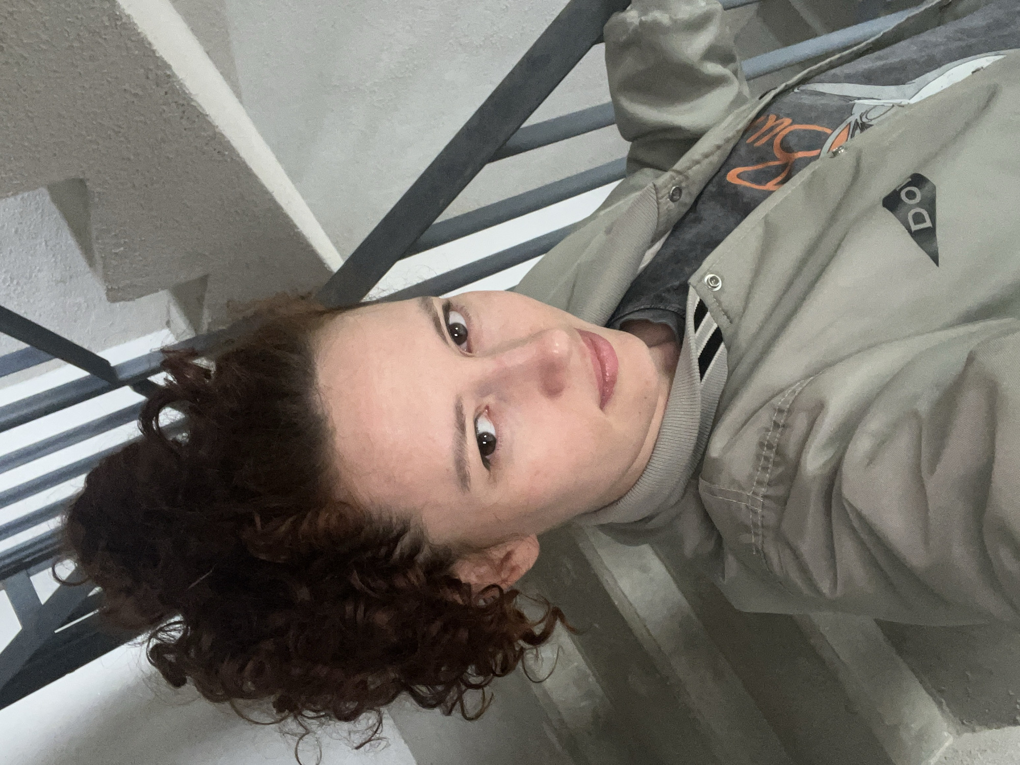 A person with brown curly hair is sitting on the steps of a staircase in Saint Petersburg, Russia. The person is wearing a gray t-shirt with an orange graphic design under a light green bomber jacket. The gray staircase is in the background, and the person is leaning against the railing.