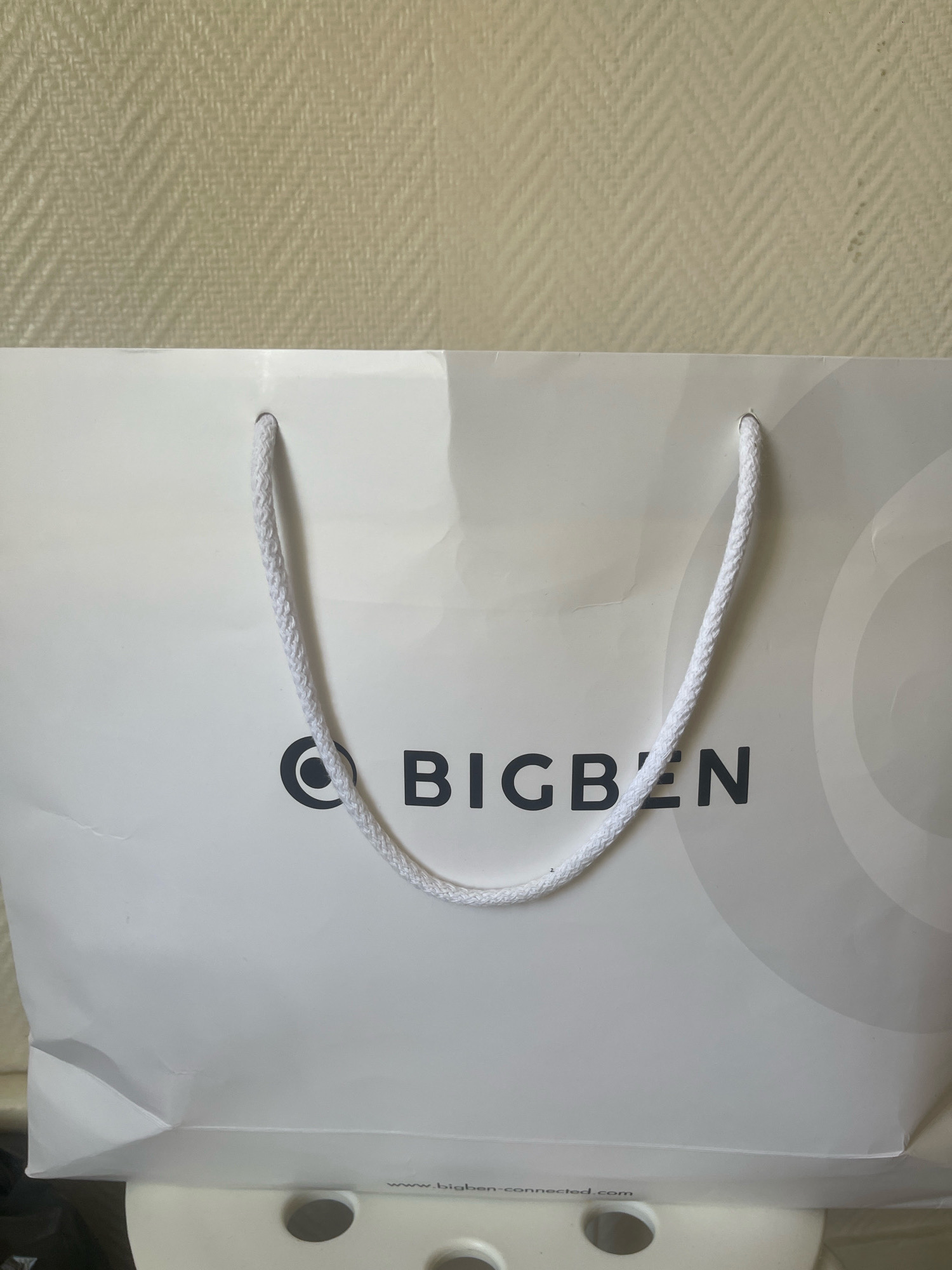 The image shows a white paper shopping bag with the logo "BIGBEN" printed on it in black. The bag has white rope handles. A website address, "www.bigben-connected.com", is printed in small text at the bottom of the bag. The bag is placed against a textured, light-colored wall. The bottom of the bag rests on a white surface with circular cutouts, possibly a piece of furniture. There are no people or other objects clearly visible in the image. The scene is indoors and appears to be a well-lit environment. The image does not provide any information about the location in the city of Paris, France.