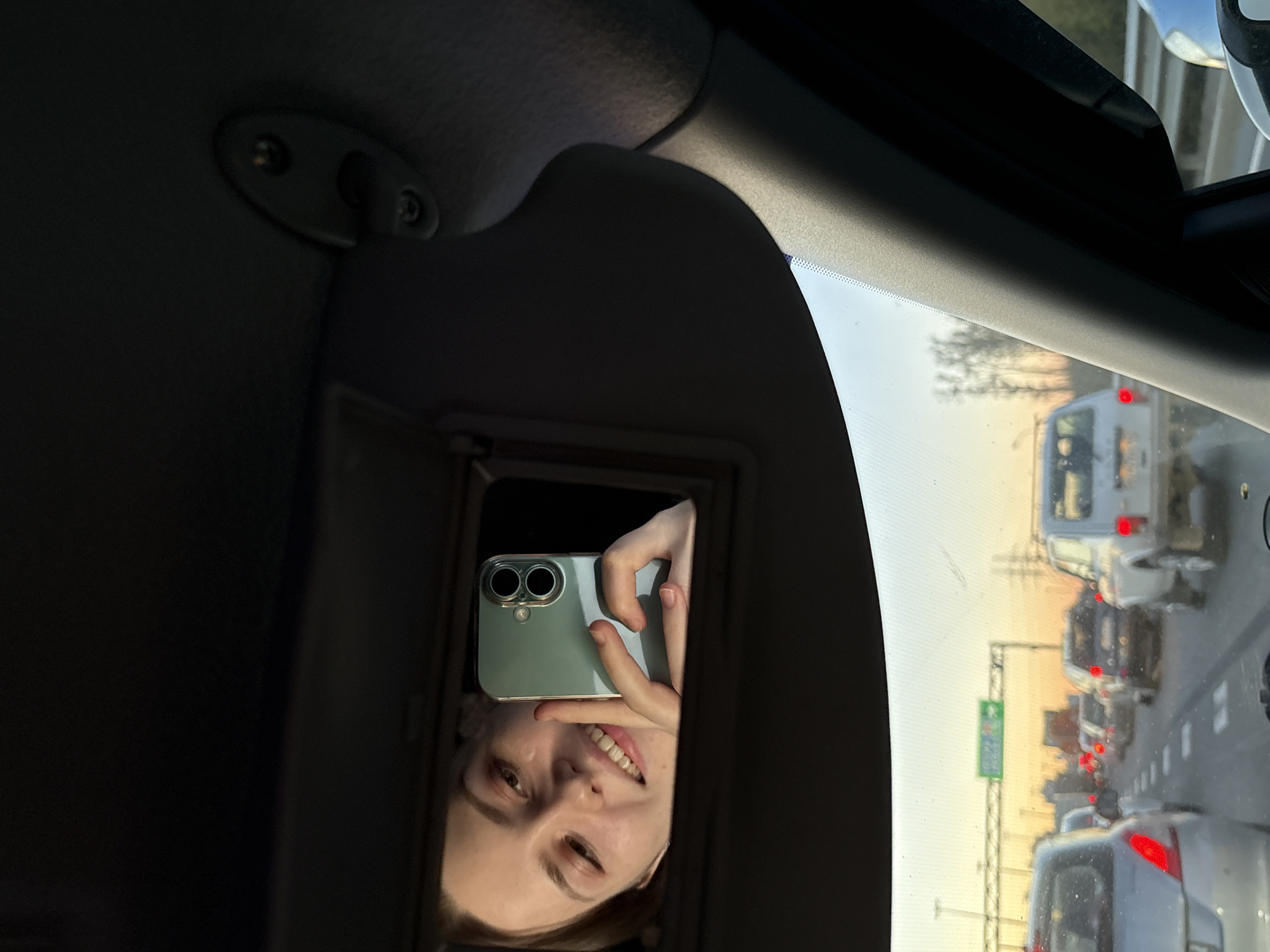 The image shows a woman taking a selfie in the mirror of a car's sun visor. She is holding a light green smartphone with two lenses. The woman has fair skin, light hair, and is smiling slightly. The car is in traffic on a road with multiple lanes. Other cars and a van are visible ahead, their brake lights illuminated. A road sign is also visible. The city is specified as Unknown, Russia. The interior of the car is visible, including the sun visor, the car's roof, and the windshield.