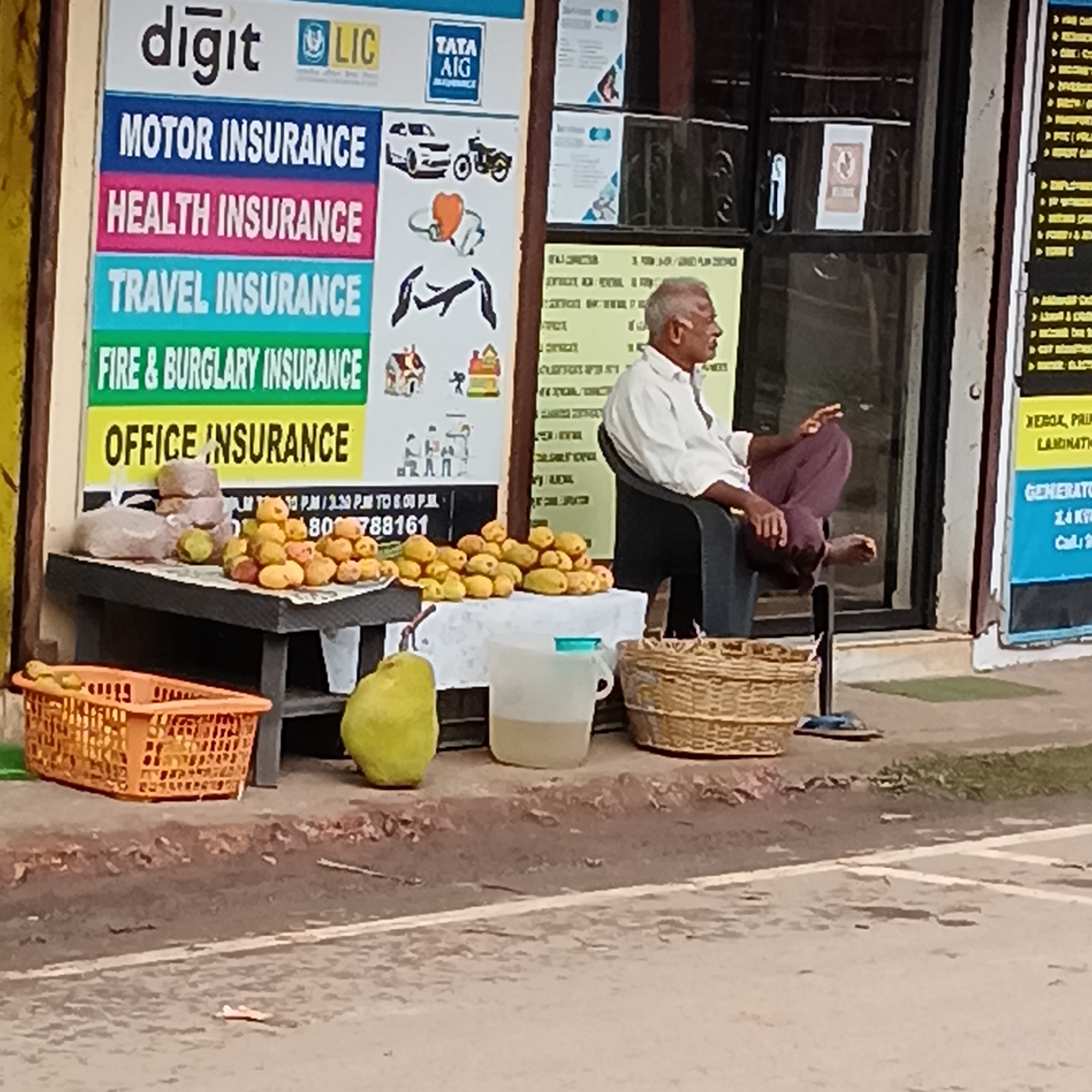 An elderly man is sitting in a black chair with a black frame, resting his bare foot on a black stand in front of a building on a street in Oxel, India. He is wearing a light-colored long-sleeved shirt and maroon pants. His hair is gray and receding. The building is next to a sidewalk. To the man's left is a makeshift stand displaying fruit, some of which are contained in baskets. Behind the fruit stand is an advertising board with insurance information for companies like Digit, LIC, and Tata AIG. There is another sign on the right side, but it is mostly cut off in the frame.