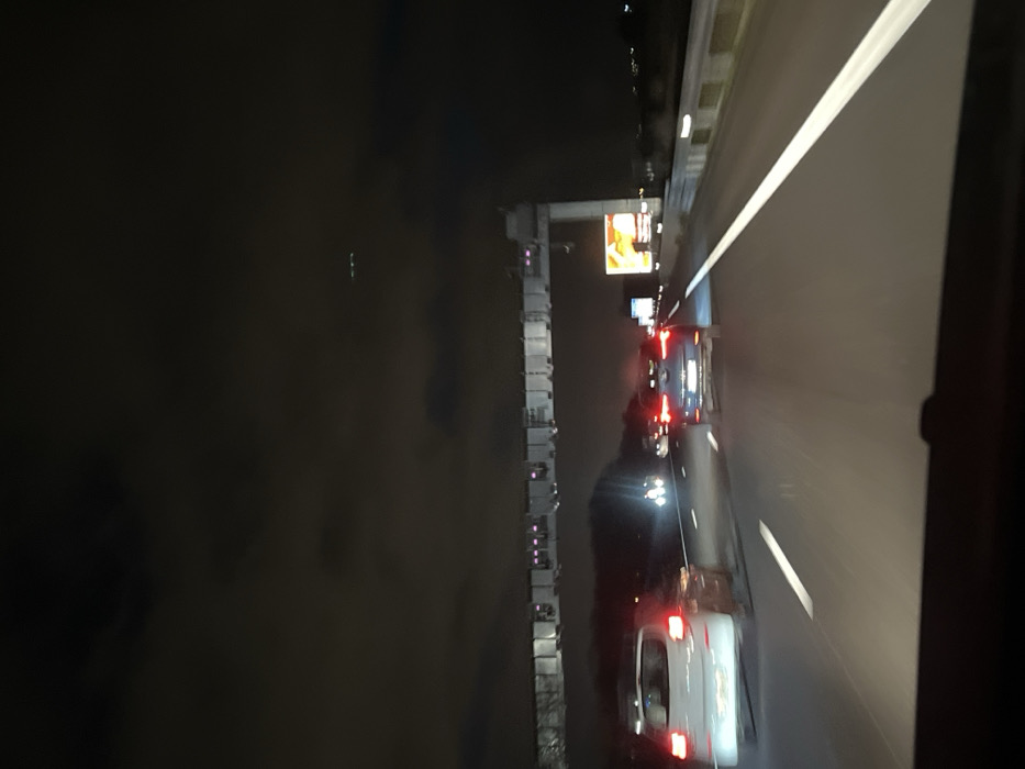 Night scene on a highway with cars driving in both directions. A bridge runs parallel with the highway, partially obscured by darkness. Advertisements are visible above the bridge. Car headlights and taillights illuminate the road. The image is taken from inside a vehicle, showing part of the vehicle's interior on the right side.