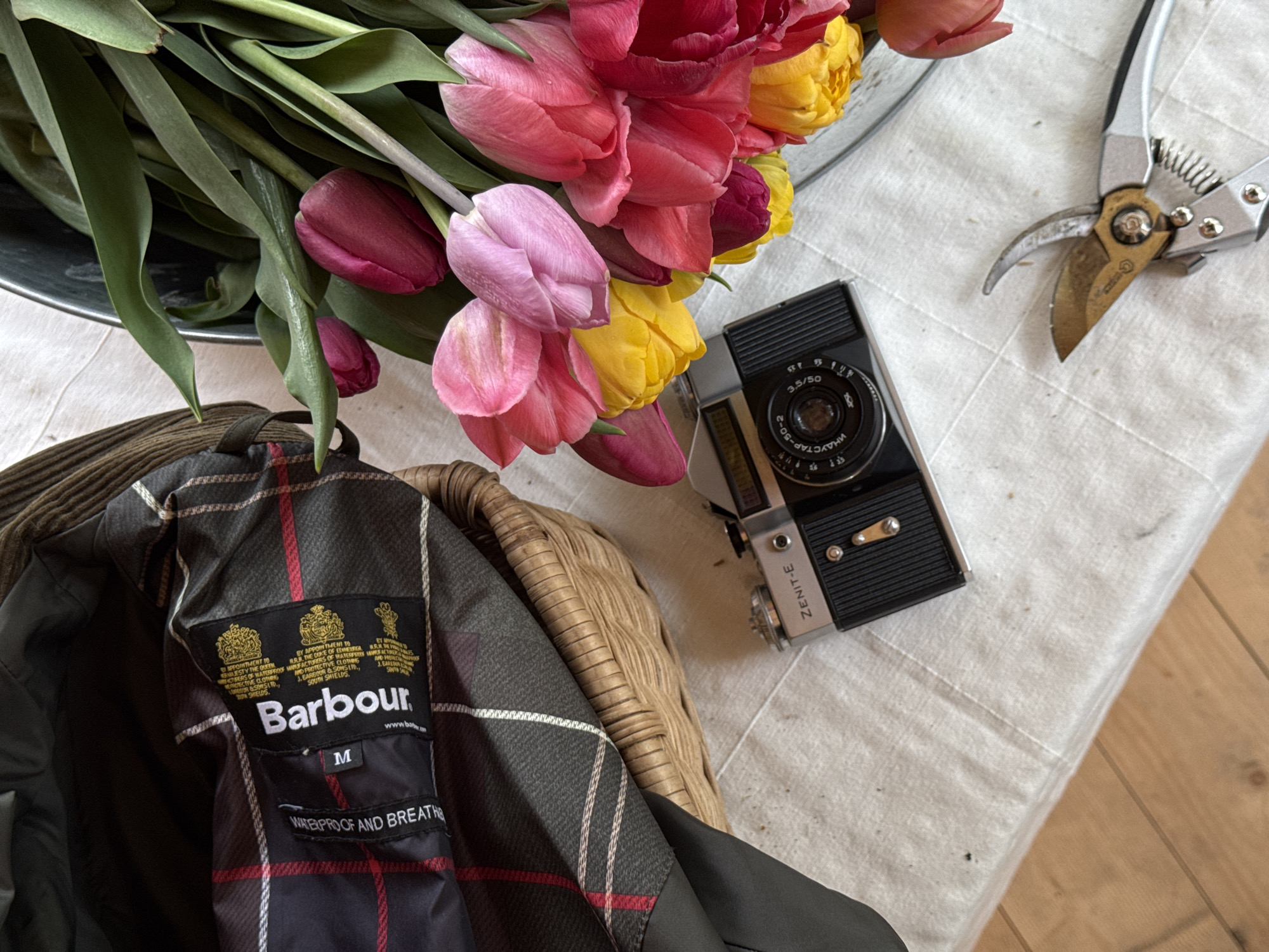The image depicts a still life on a white tablecloth with a wooden floor visible in the lower right corner. A bouquet of tulips in various colors (red, pink, and yellow) sits in a metallic container. Next to the tulips, there's a vintage Zenit-E camera and a pair of garden shears. Partially visible is a Barbour jacket with its label clearly displayed. The jacket's lining features a plaid pattern with red and white lines. The Barbour label indicates a size M and mentions "WATERPROOF AND BREATHABLE." No information in this image indicates its location is in Sapronovo, Russia.