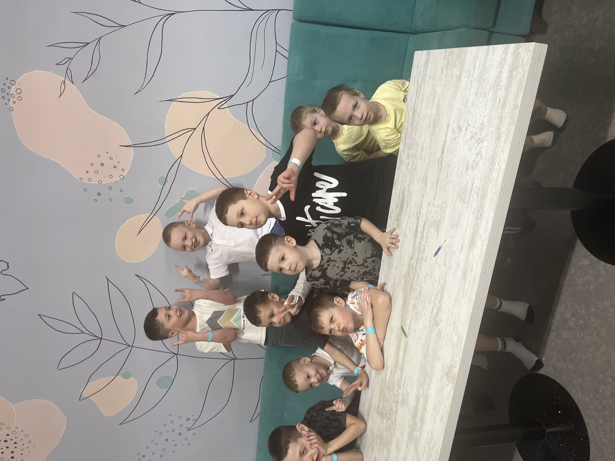 A group of nine boys are gathered around a light-colored table in a room. The boys are of varying heights and are wearing different colored shirts, including yellow, white, black, and gray. Some of them are making hand gestures, and all are looking toward the camera. The background features a wall with a gray base color and a design of abstract shapes and lines resembling plants. A green cushioned bench is behind some of the boys. The floor is gray. The setting is a building in Ulyanovsk, Russia.
