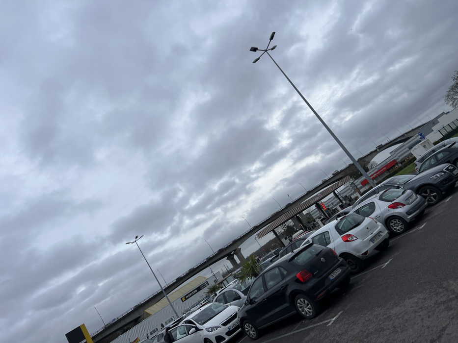 This image depicts an outdoor parking lot under an overcast sky, taken with a pronounced tilt from the bottom-left to the top-right. The location is identified as Hérouville-Saint-Clair, France, specifically in a commercial area near an elevated road.
In the foreground and midground, numerous passenger vehicles are parked in rows on an asphalt surface. The vehicles include various hatchbacks and sedans, predominantly in white, black, and silver/grey colors. A white Peugeot 208 is visible on the far left, followed by a black Volkswagen Polo, and several other compact cars. Some vehicles have French license plates visible (e.g., ES-554-CL on the black Polo, ES-854-HX on a white hatchback). The asphalt appears damp in areas.
Two tall, grey lampposts with multiple light fixtures (four per pole, arranged radially) extend upwards. The one in the far right midground is particularly prominent, reaching towards the upper right corner of the frame.
In the background, an elevated road or bridge, supported by grey concrete pillars, traverses horizontally across the scene. Below the elevated road and behind the parked cars, several commercial buildings are visible. A yellow sign for "NORISKO Auto" (a French vehicle inspection network) is prominent on the left. Further to the right, a fuel station canopy with red signage and several pump islands is identifiable. A digital display at one of the pumps shows red numbers, possibly indicating fuel prices (e.g., "1.898"). Beyond the fuel station, a building with a red facade and a white sign partially visible as "LE PREM" is seen. Sparse vegetation, including a palm-like plant and bare trees, is present around the commercial structures.
The sky is entirely covered with dense, grey clouds, indicating an overcast day. No individuals are visible in the scene, and no specific actions or interactions are depicted. The overall impression is one of a typical commercial parking area on a dull, possibly damp, day.