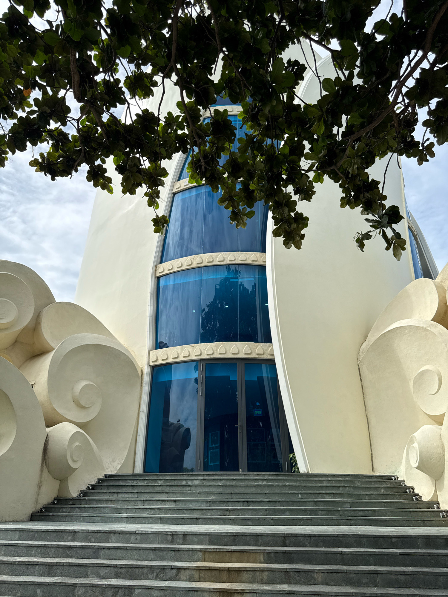 This is a low-angle, medium shot of the entrance to the Nha Trang Institute of Oceanography in Nha Trang, Vietnam. A large tree with dark green leaves hangs into the frame from the top, partially obscuring the sky and the building's upper portion. The building itself is a cream-colored, modern structure with a curved facade. On either side of the central entrance are decorative, swirling white elements that resemble stylized clouds or waves. A wide set of stone stairs leads up to the entrance, which features a large glass door with a reflective blue tint. The sky is overcast with patches of bright white clouds.