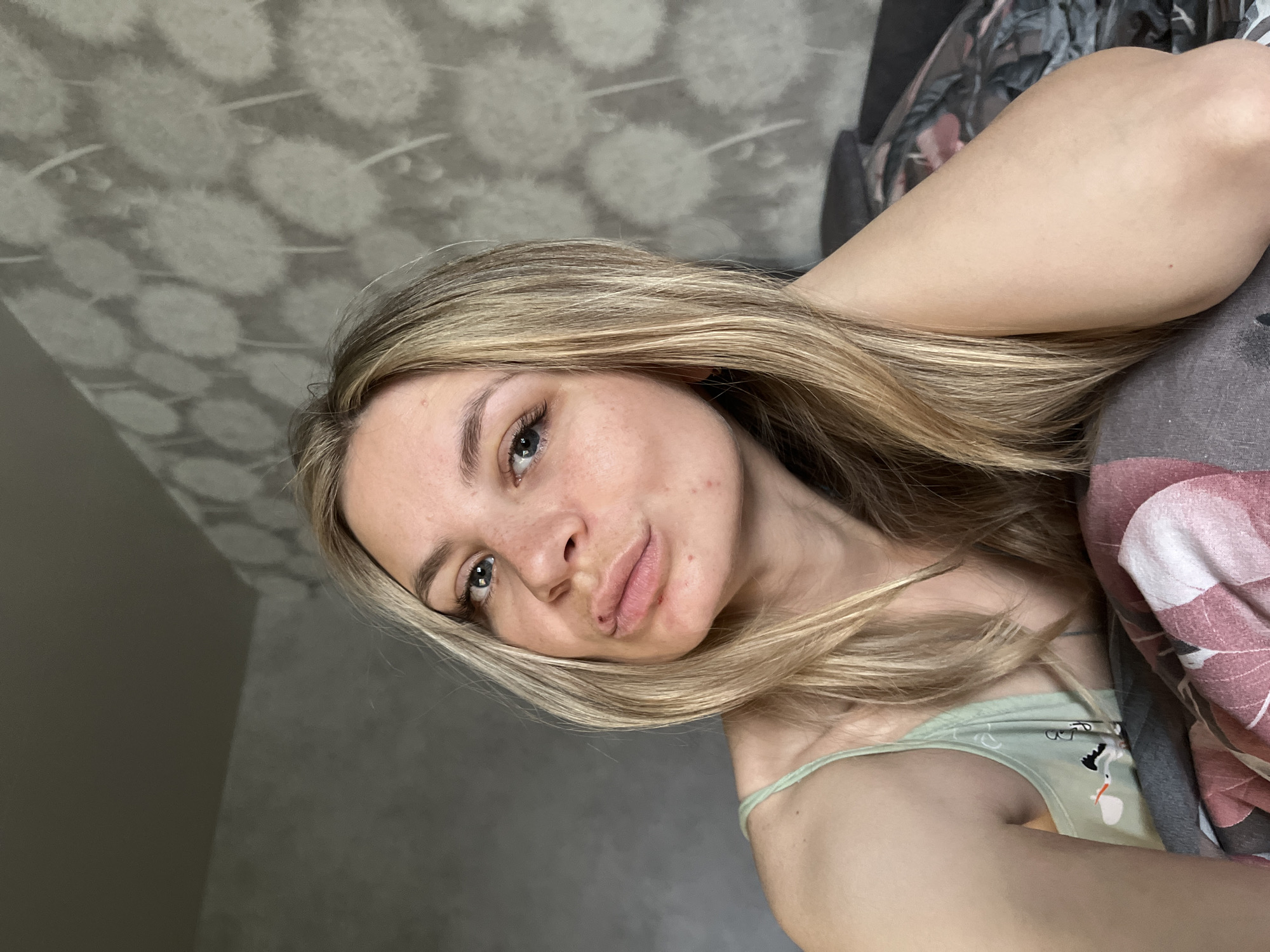 A young woman with blonde hair lies on a bed in what appears to be a bedroom. She is wearing a light green top with a design on it, partially obscured by a pink and grey blanket. The wall behind her is decorated with a grey and white dandelion-like pattern. The location is identified as being in the city of Chaikovsky, Russia, based on the available information.