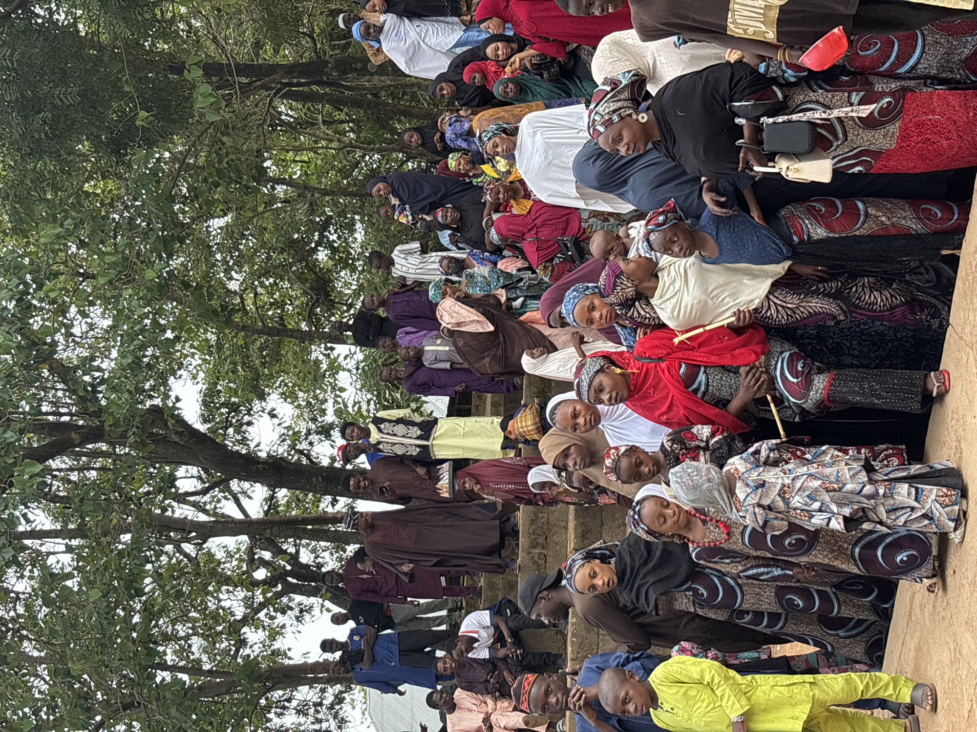 The image captures a gathering of people, likely a community event or celebration, in an outdoor setting with trees providing shade. The scene appears to be on a sloped area with multiple tiers of concrete steps where many individuals are seated or standing. The majority of the attendees are dressed in traditional African attire, with vibrant colors and patterns visible in their clothing and headscarries.
In the foreground, a young boy in a bright yellow outfit stands on a dirt path, facing left. Beside him, two young girls in patterned dresses are looking downwards. Further back, a crowd of people are seen on the steps, with men and women of various ages present. Some individuals are engaged in conversation, while others appear to be observing something. The atmosphere seems festive, suggested by the attire and the gathering itself.
The environment consists of lush green trees with large leaves, indicating a possibly humid or tropical climate. The lighting suggests it might be daytime, possibly overcast or under dappled sunlight. There are no explicit signs or landmarks to pinpoint the exact location within Jos, Nigeria, but the overall visual elements are consistent with a rural or semi-urban setting in such a region. The image is taken from a slightly elevated perspective, looking down at some of the people on the steps.