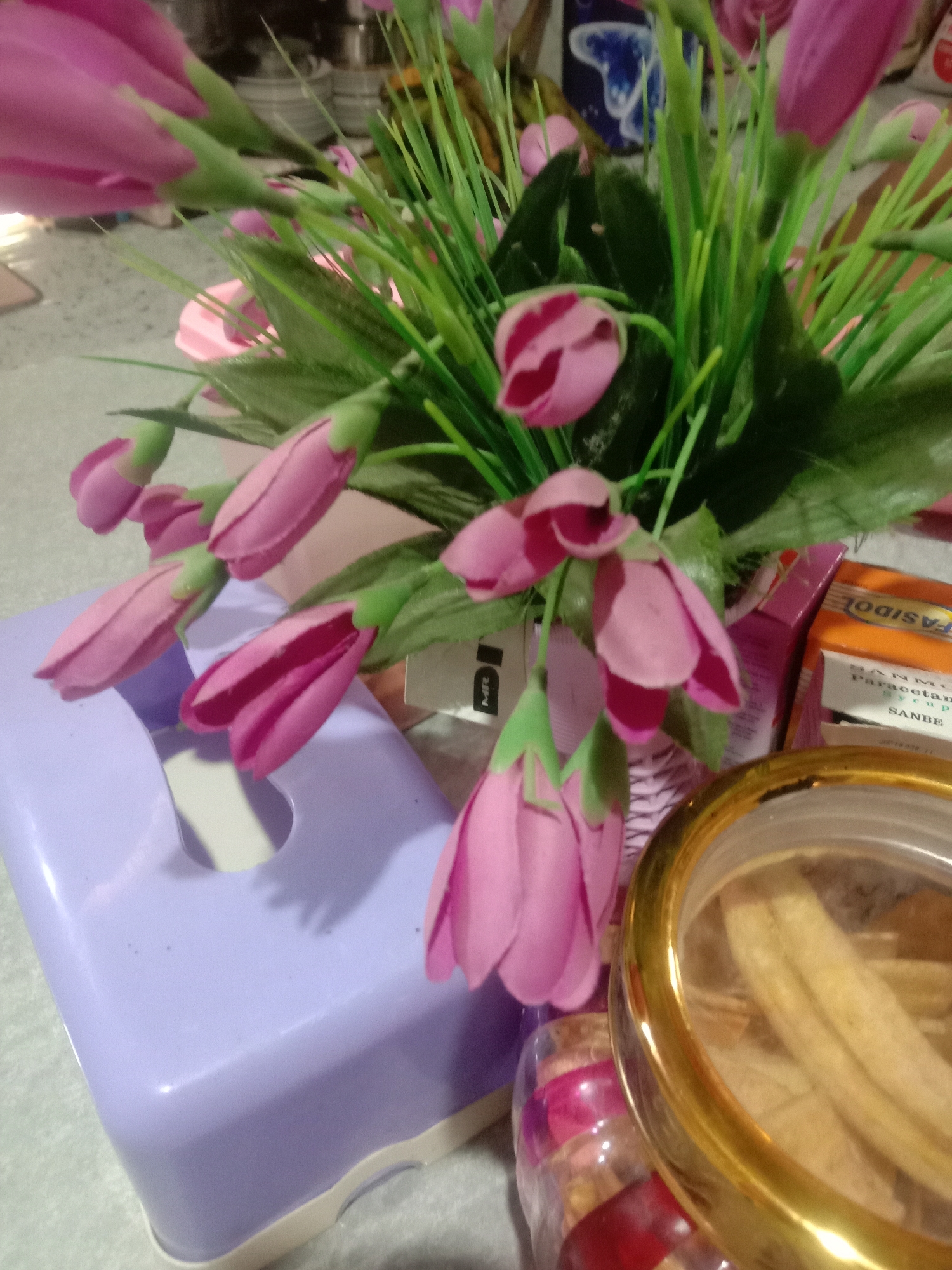 The image shows a close-up of a collection of items on a surface, seemingly indoors. There is a vase of artificial pink tulip-like flowers with green stems and leaves. Adjacent to the flowers is a light purple plastic tissue box. Next to the flower arrangement is a box of "FASIDOL" paracetamol syrup, and a transparent jar with a gold-colored lid, containing pale, stick-like objects.
There is no information in the image indicating a specific location in the city of Lhokjok, Indonesia.