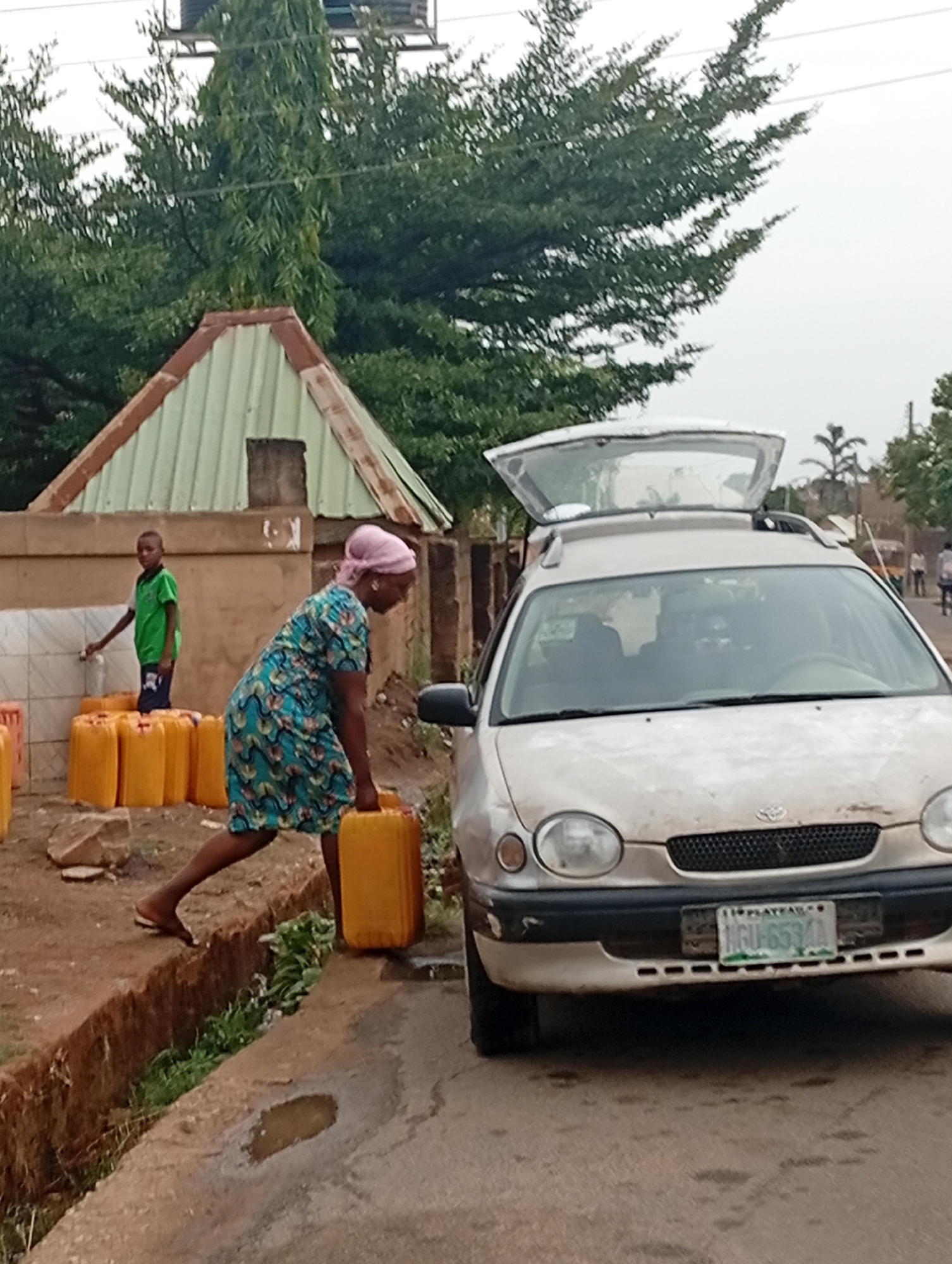 The image depicts a woman in a blue dress and pink headscarf carrying a yellow jerrycan near a car with its trunk open on a street in Jos, Nigeria. A young boy in a green shirt is standing by a water source with several yellow jerrycans lined up nearby. The location appears to be a residential area, with a small building having a peaked roof visible in the background and trees surrounding the area. The car, with license plate "MGU-653AA Plateau," is parked on the side of the road. The overall scene suggests water collection is taking place.