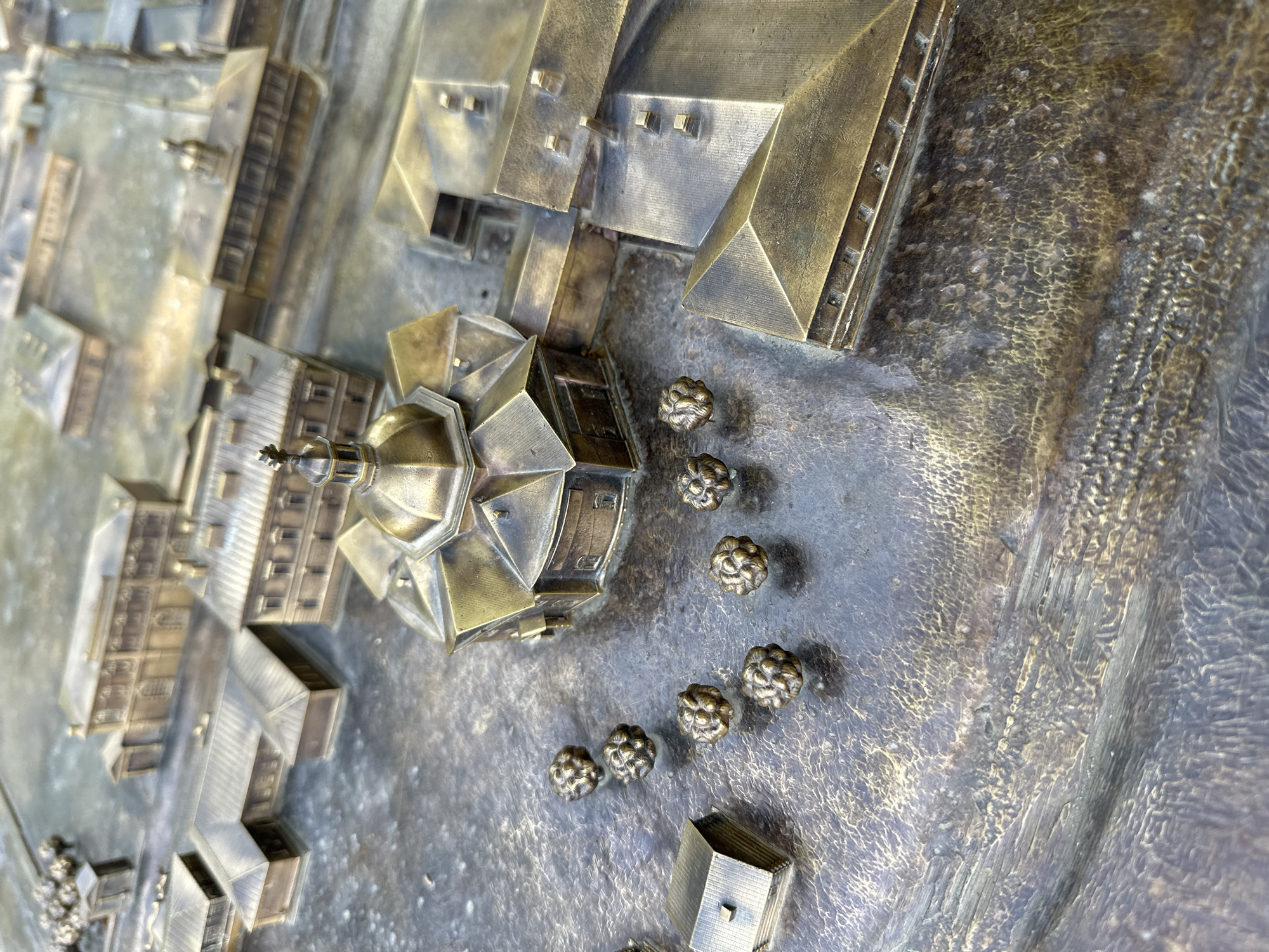 This is a close-up, high-angle, eye-level shot of a bronze architectural model. The model depicts a city with multiple buildings and a central circular structure topped with a dome and a spire. The buildings are intricately detailed, showing roofs, windows, and entrances. The ground surface of the model is textured, resembling cobblestones or uneven terrain. Scattered across the ground are several small, spherical objects that appear to be stylized trees or bushes. In the lower left quadrant of the image, a small, rectangular building with a pitched roof is visible. The lighting is natural, casting subtle shadows and highlighting the metallic sheen of the bronze. The model is located in Barnaul, Russia, and represents the area surrounding the Barnaul Ascension Cathedral.