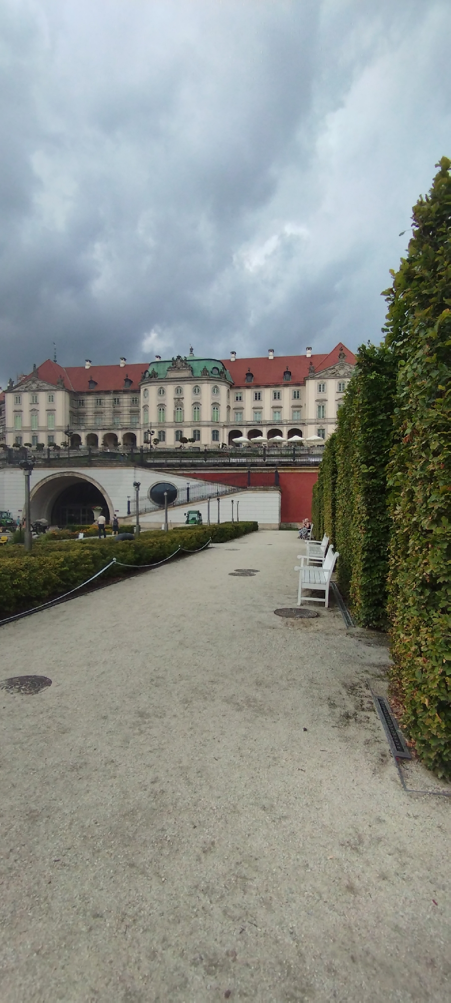 The image depicts a view of the Royal Castle in Warsaw, Poland, from the gardens. The castle, with its red-tiled roof and pale facade, is a prominent structure in the background. In front of the castle, there is a red wall and a terrace with white umbrellas. The foreground features a gravel path lined with manicured green bushes on both sides. Two white benches are visible on the right side of the path, and a person is seated on one of them. The sky is overcast with dark, heavy clouds, suggesting an impending storm. The overall scene is one of a historical landmark in a well-maintained garden setting, under a moody sky.