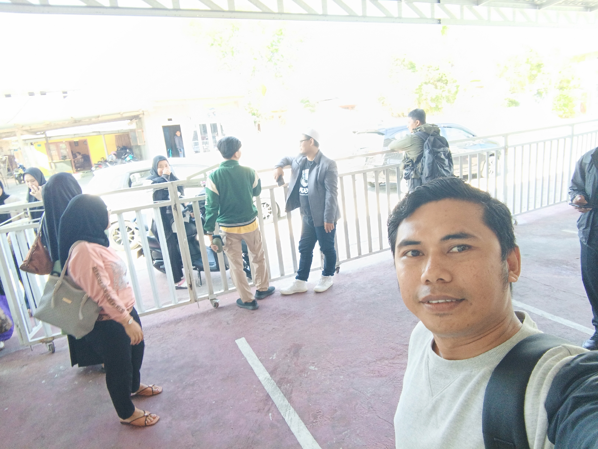 The image shows a group of people standing behind a white fence, possibly at an outdoor gathering or waiting area. In the foreground, a man is taking a selfie, looking directly at the camera. He is wearing a light grey sweatshirt and a backpack. Behind him, to his left, a woman wearing a black hijab and a pink long-sleeved shirt is visible, carrying a grey bag. Other individuals, some wearing hijabs and others in more casual attire, are dispersed behind the fence. One person in a green jacket and khaki pants is looking towards the right of the frame, while another in a grey suit jacket is leaning on the fence, holding a phone. A man in a dark jacket with a backpack is also present. The environment appears to be outdoors, with sunlight filtering through a covered structure above. There are buildings and some vegetation visible in the background, suggesting an urban or semi-urban setting. The ground is paved with a reddish-pink surface marked with a white stripe. The overall atmosphere seems casual, with people interacting or waiting. The presence of buildings with open structures and a paved ground suggests a public space, possibly an event venue or a transit point within the city of Gorontalo, Indonesia.