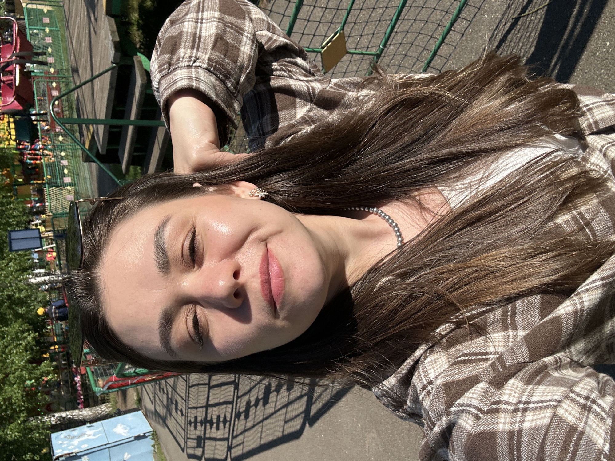A woman with long brown hair is posing for a selfie outdoors. She is wearing a brown and white plaid shirt, a white top, and a silver beaded necklace. Sunglasses are perched on top of her head. Her eyes are closed and she is smiling slightly. The background includes a green metal fence, a playground or amusement park ride with red seats, trees, and a blue structure. The location is possibly in the city of Chaikovsky, Russia. The ground is paved and dappled with shadows.