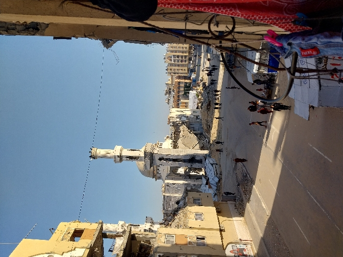 The image presents a ground-level view of a significantly damaged urban landscape in Khan Yunis, Palestinian Territory, under a clear blue sky. The central feature is the Great Mosque of Khan Yunis, or Al-Kabir Mosque, with its minaret largely intact but the main mosque structure behind it extensively damaged, featuring a partially collapsed dome and walls, exposing internal rubble and structural elements.
To the left, a multi-story building exhibits severe structural damage, including exposed rebar and missing wall sections. Other buildings in the mid-ground and background also show varying degrees of destruction. The foreground features a paved street with faded white lane markings, largely clear of major debris but with scattered smaller rubble along its edges.
Numerous individuals are observed walking across the street and through the damaged areas. A person in a long, dark red garment walks centrally in the street. On the right side of the street, a cluster of approximately 8-10 people gathers near several small, makeshift market stalls or shelters, some with blue tarps. One individual in a dark uniform is visible within this group.
Long shadows are cast across the street from buildings on the left, suggesting morning or late afternoon light. In the immediate right foreground, elements of a building's exterior, including a railing and clothes hanging to dry (a red patterned cloth, blue jeans, and other garments), are visible, indicating the vantage point of the photographer. Overhead, a thin, dark utility line spans the sky.