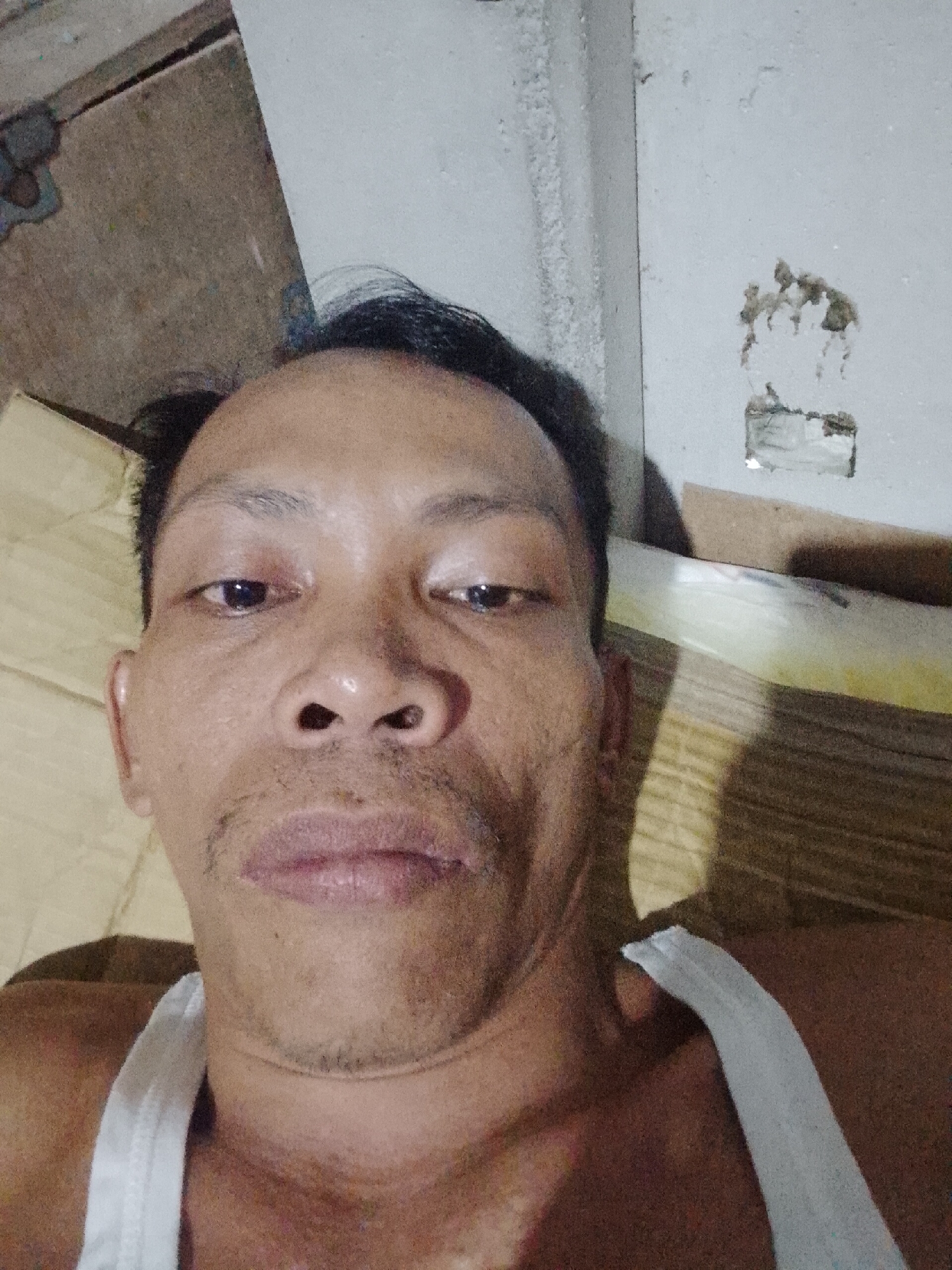 The image is a selfie of a man with dark hair and brown skin, wearing a white tank top. He appears to be lying down, possibly on a mattress or bedding. The background consists of a wall with damaged paint or paper, and a wooden surface. The location could possibly be the city of Bukit Ketok, Indonesia.