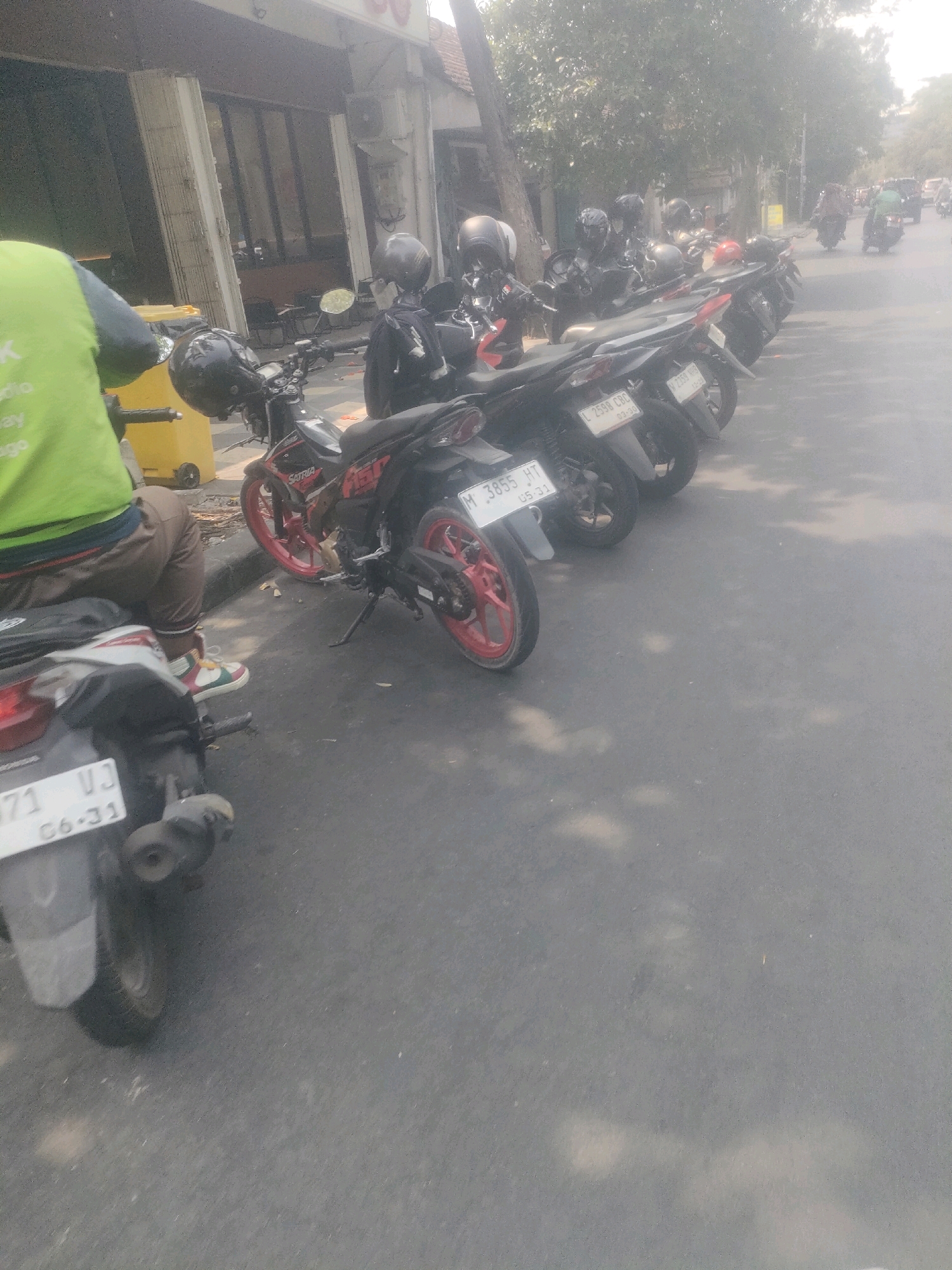 The image depicts a street scene in Surabaya, Indonesia, with a row of parked motorcycles on the right side of the frame. In the foreground, a person wearing a bright green vest and brown pants is seated on a motorcycle, facing away from the camera. The person is wearing colorful sneakers. To the left of the person is a yellow trash bin. The motorcycles are parked along the curb of a paved street. The motorcycles themselves are varied in color, with some having red rims. License plates are visible on some of the motorcycles, with markings such as "M 3855 HT" and "L 2598 CBC". In the background, there are buildings and trees, suggesting an urban environment. Sunlight filters through the trees, casting shadows on the street. Further down the street, other motorcycles and individuals can be seen, with traffic appearing to be moving in the distance. The overall atmosphere is that of a typical day in a bustling Indonesian city.