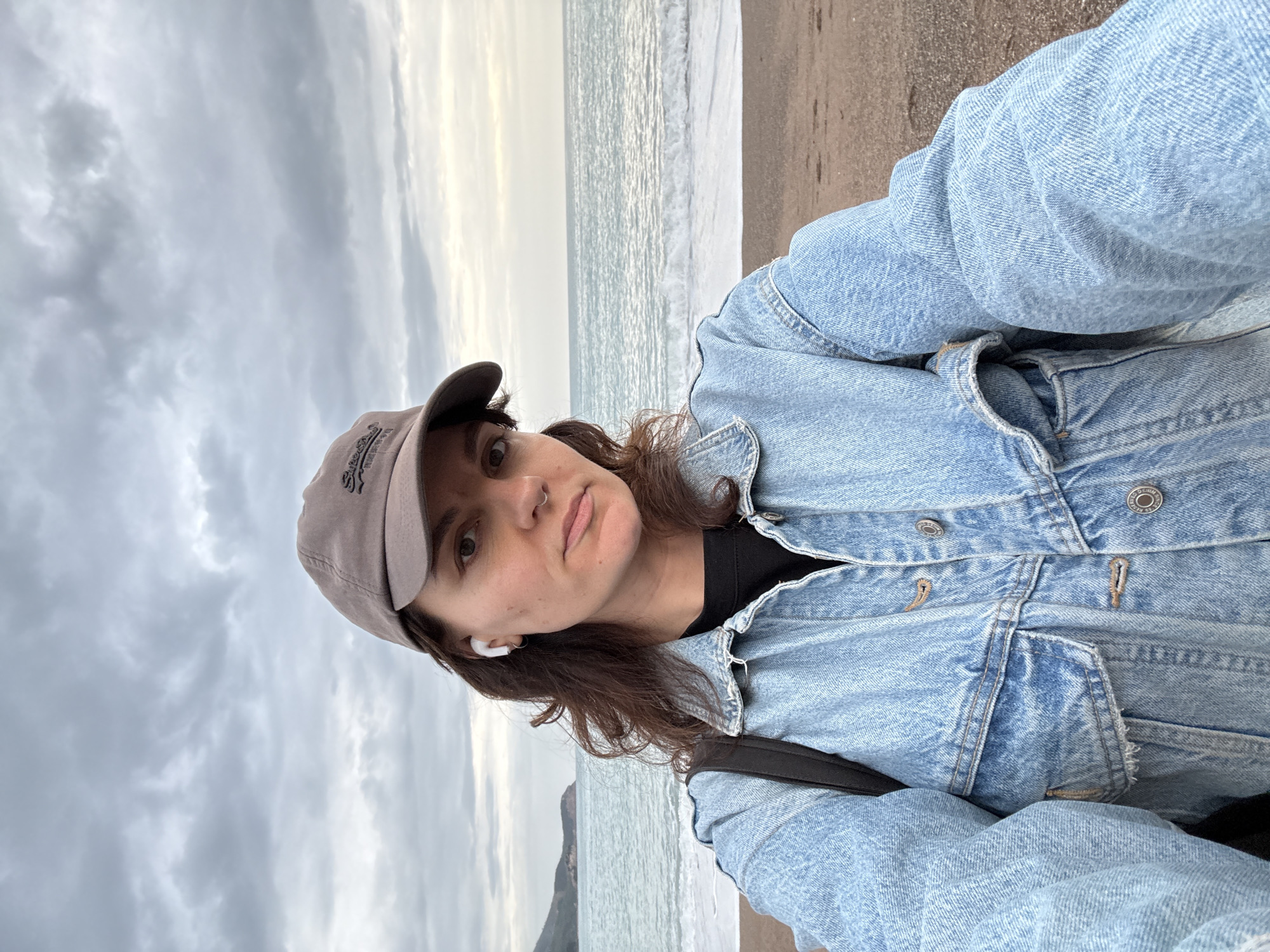 A vertical, medium-shot selfie of a person wearing a denim jacket and a baseball cap, taken outdoors. The person is positioned on a beach, with the sea and cloudy sky in the background. The beach appears sandy and dark. The sea has small waves. A land mass appears on the horizon. The person has brown hair and is wearing a white earbud. The person also has a nose ring. This beach is in Rafailovići, Montenegro.