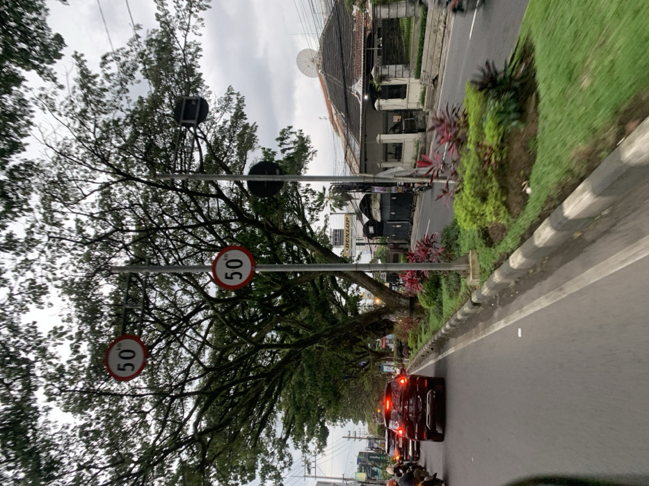 The image shows a street scene in Malang, Indonesia, located at latitude -7.94 and longitude 112.637. A black SUV is traveling on the road in the middle of the frame. There is a row of trees on the left side of the road, and a curb with green grass and bushes. Two round speed limit signs reading "50" are visible on a pole on the left side of the image. A building with a white facade is visible in the background. The road surface is black asphalt with white markings. The sky is cloudy, and the scene is captured during daytime. A motorcycle is visible in the far right corner of the image, traveling in the opposite direction of the SUV.