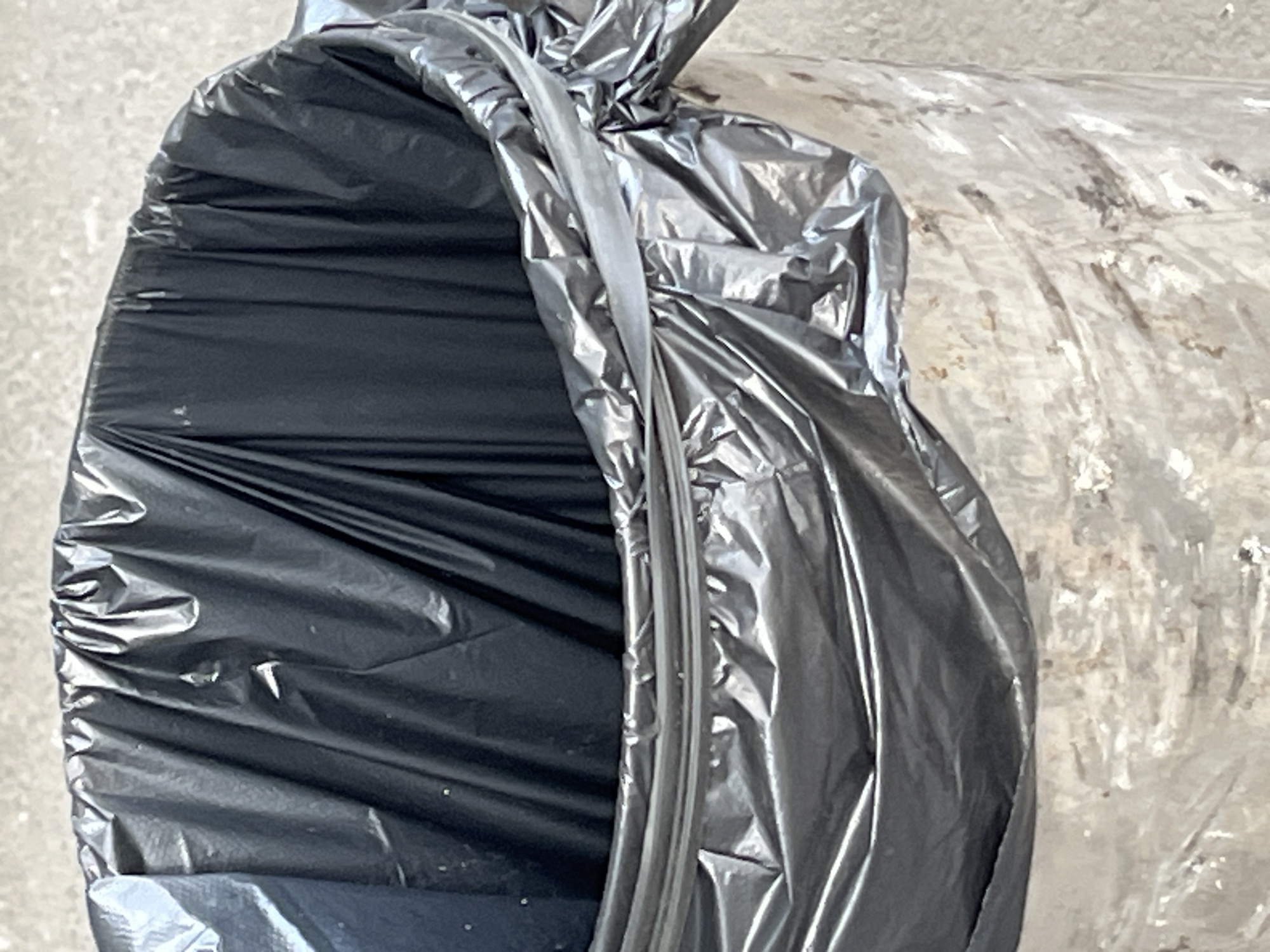 This is a close-up, overhead shot of two large, cylindrical objects wrapped in plastic. The object on the left is a black plastic bag, its opening partially unzipped, revealing a crumpled interior. The bag is secured with a gray, ribbed zipper. To the right of the black bag, a second object is visible, wrapped in a translucent, crinkled plastic material. This material appears to be slightly dirty and has an uneven texture, with darker smudges and lighter, opaque patches. The background is a rough, gray, concrete-like surface, suggesting an outdoor or industrial setting. The lighting is even and diffused, with no strong shadows, indicating it might be overcast or the light source is not directly visible. The focus is sharp on the plastic wrappings. There are no people or text visible in the image. The scene appears to be a still life of protected objects, possibly awaiting transport or storage. The context of Kyiv, Ukraine, does not provide any specific visual cues within the image itself.