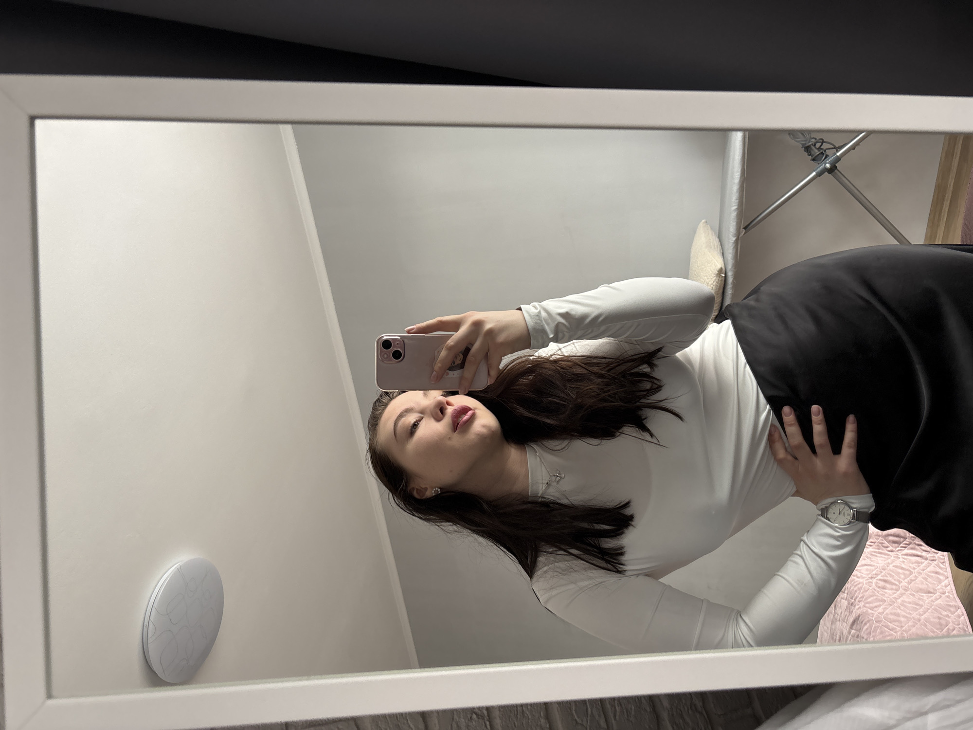 A young woman takes a selfie in a mirror. She is wearing a long-sleeved white shirt and a black skirt. She is holding a pink smartphone in her left hand. She has long, dark hair. The mirror is white and rectangular. The background is a white wall with a light fixture and a clothes drying rack. The location is most likely in a residential area of Perm, Russia.