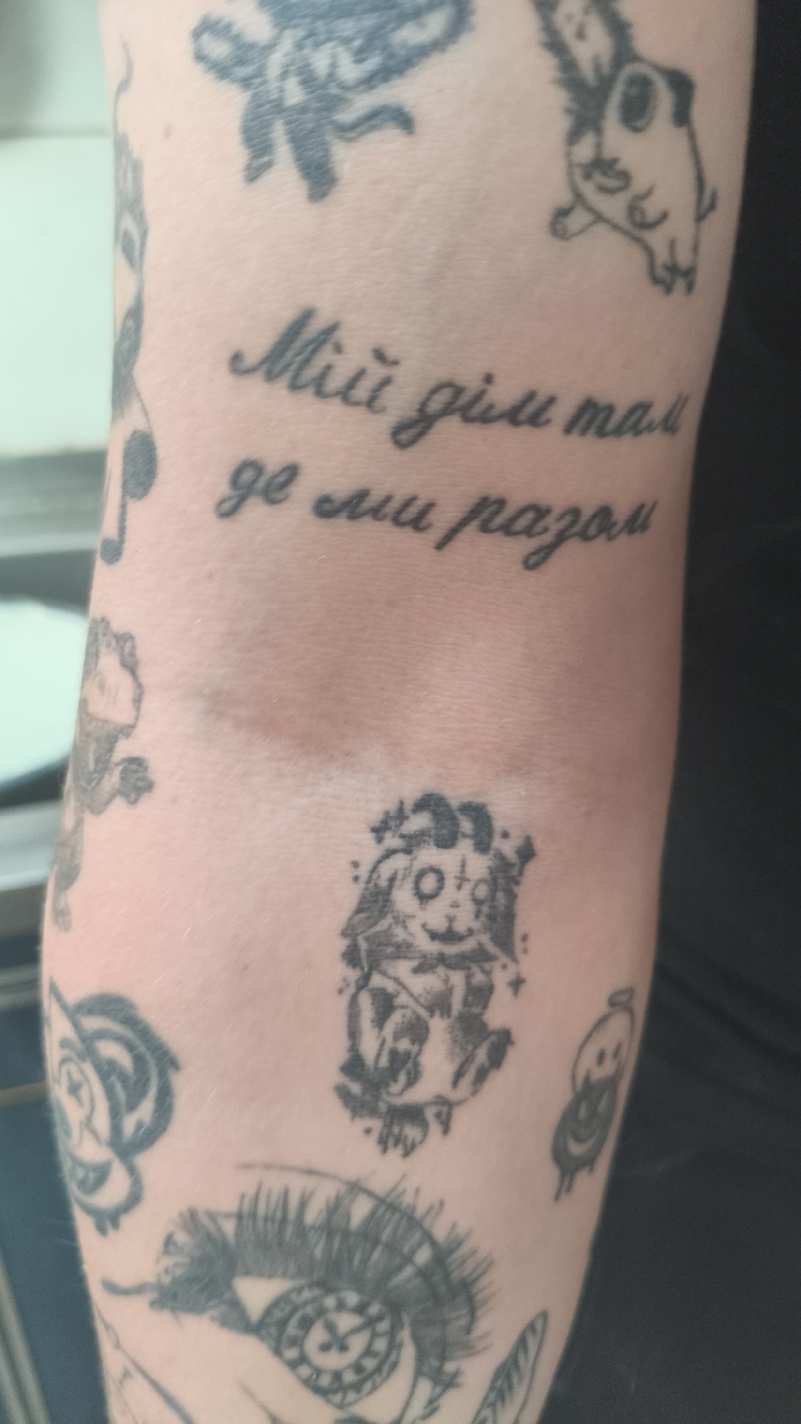 The image shows a close-up of a person's arm covered in various tattoos. The tattoos include cartoonish figures, an eye design, and text in a script that appears to be Slavic. The text reads "Miii gim mall ge mu pazon". The tattoos appear to be black ink. The background is out of focus and indistinct, revealing no information about Nowy Tomyśl, Poland.