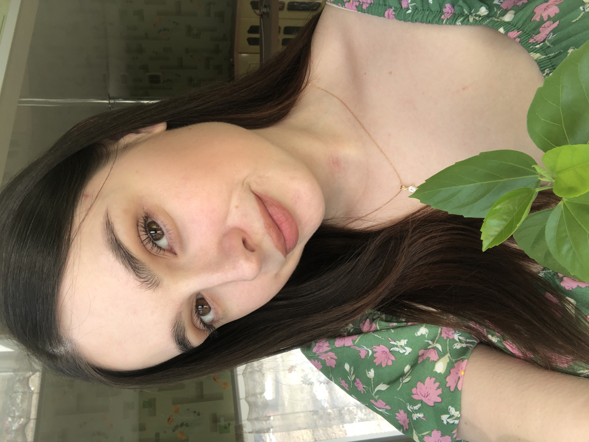 A portrait of a woman with fair skin and brown hair is angled towards the viewer. She has brown eyes, distinct eyebrows, and is wearing a light lipstick. A delicate gold necklace with a small pendant is visible around her neck. The woman is wearing a green floral-patterned dress. To her right, some green leaves are partially obscuring her shoulder. The background is out of focus, but suggests an interior space. The location is unknown and cannot be determined to be Noviy, Russia from this image.