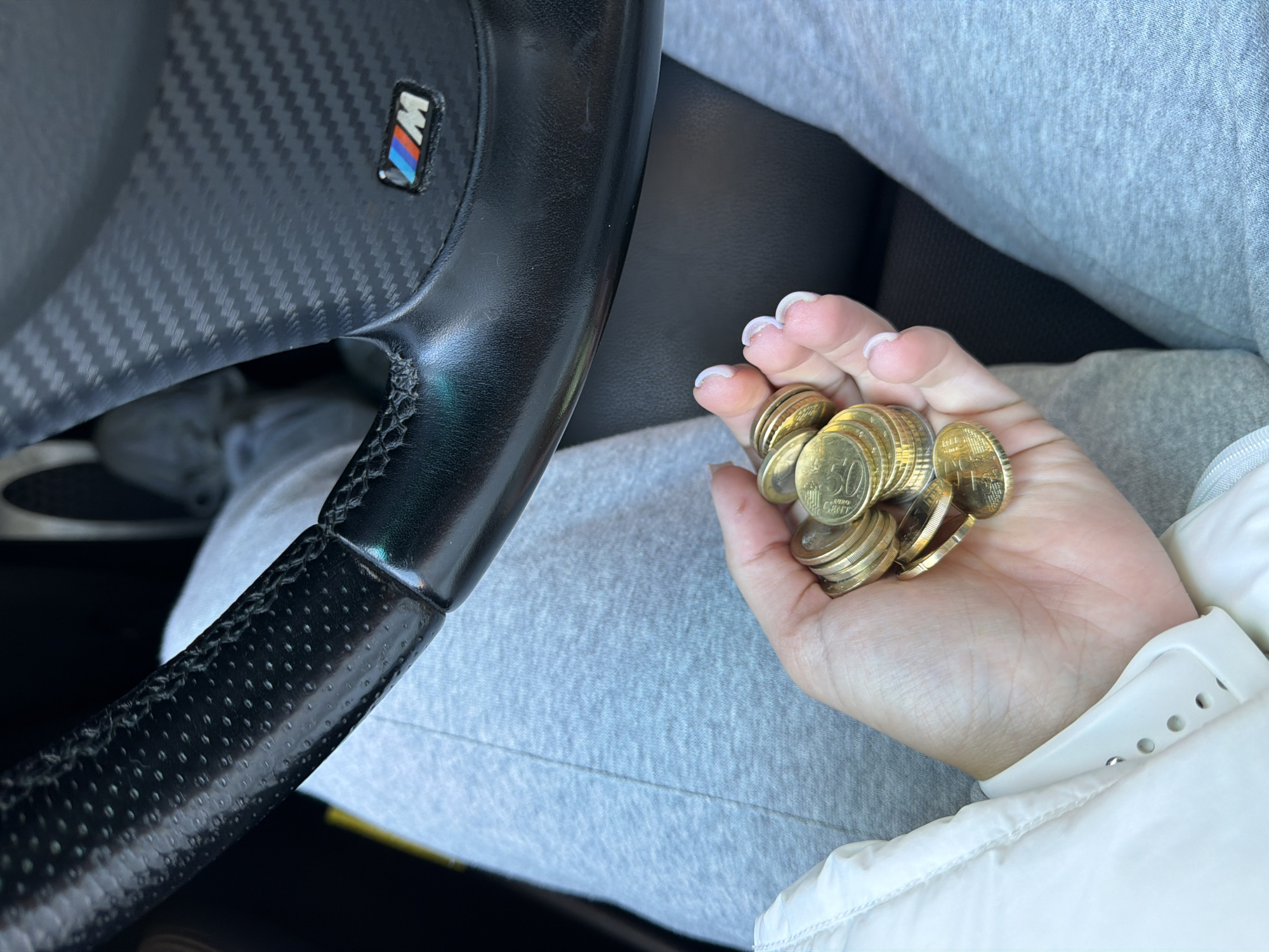 The image shows a person in the driver's seat of a car, holding a handful of Euro coins. The steering wheel has a carbon fiber design with a BMW "M" logo. The person is wearing a light gray sweat suit and a white puffy jacket. The coins appear to be mostly 50 Euro cent pieces. The car interior is partially visible. Based on the coins, the location is likely in Europe. Without further information, determining the specific city of Gabrovo, Bulgaria, is not possible from this image.
