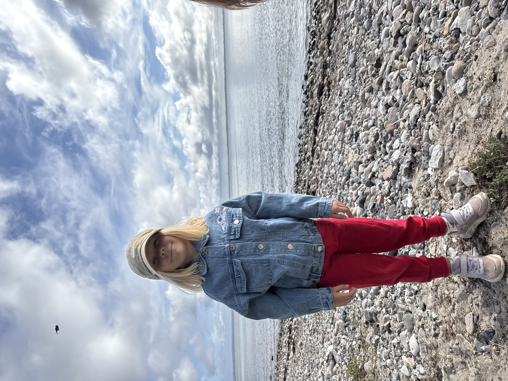 A young girl with blonde hair and a light-colored baseball cap stands on a rocky beach, facing the camera. She is wearing a denim jacket over red pants and grey sneakers. The sea is visible behind her, with a cloudy sky above. A small bird is flying in the sky. The ground is covered with pebbles and sand. The scene appears to be a coastal area.