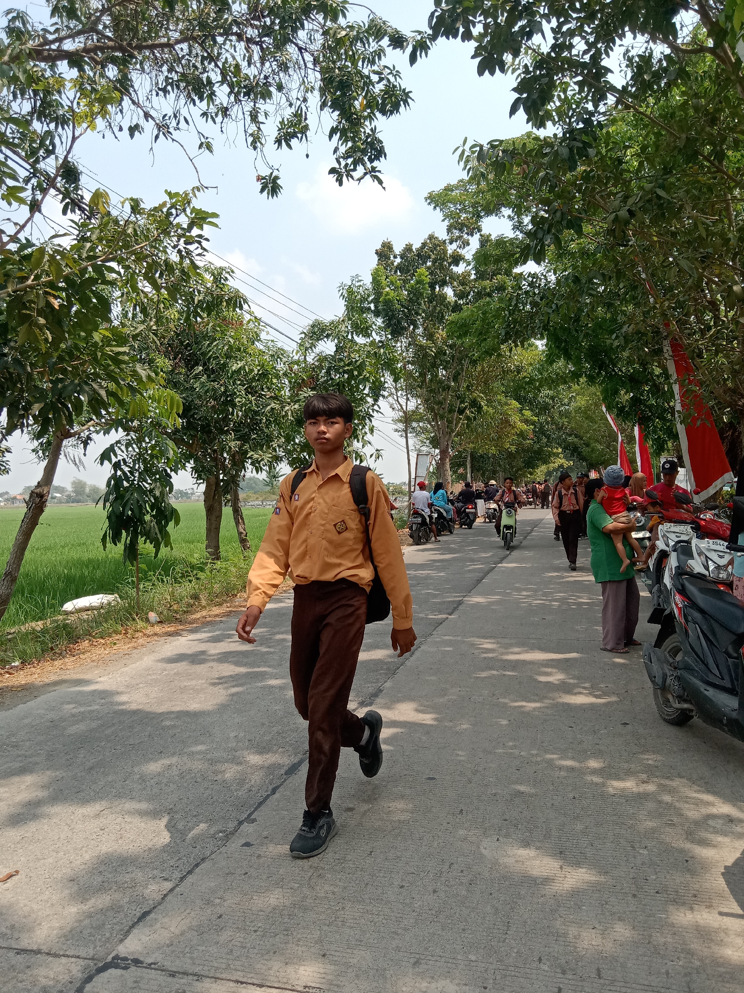 A young boy in a tan school uniform with brown pants and a black backpack walks on a paved road in Kenep, Indonesia. The road is lined with trees casting shadows on the pavement. To the left of the road is a lush green rice field. In the background, several people on motorcycles and on foot are visible, with some red and white flags decorating the scene. The sky is clear and blue with some scattered clouds.
