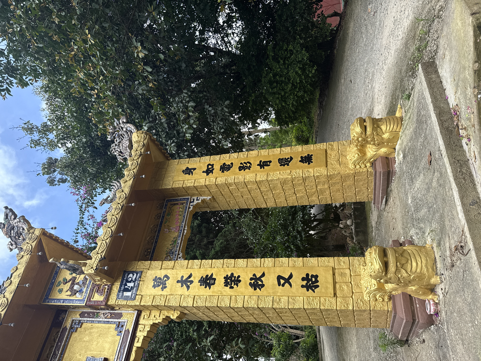 An ornate, yellow, ornate gateway stands at the entrance to a temple in the Lâm Viên area of Đà Lạt, Vietnam. The gateway is constructed of brick and adorned with intricate carvings and golden decorations. Chinese characters are inscribed on the pillars of the gateway. On the left pillar, the characters read "萬木春榮秋又枯," and on the right pillar, they read "身如電影有還無." A sign with the number "142" and Vietnamese text is visible on the top left section of the gateway. Two golden lion statues are positioned at the base of the gateway pillars, guarding the entrance. The gateway is surrounded by lush green trees and foliage, with a pathway leading through the arch. The sky above is partly cloudy. The overall scene depicts a serene and culturally rich temple entrance.