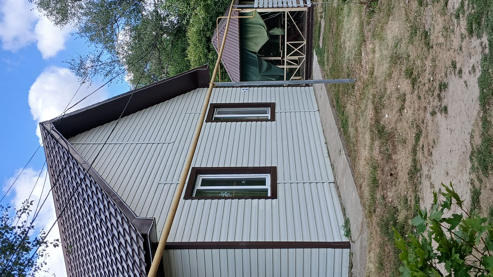 A low-angle medium shot captures a light-colored, horizontal-siding house with a brown-trimmed, dark-shingled roof. Two narrow windows with white frames and brown trim are visible on the house's side. A yellow horizontal pipe runs across the facade of the house, supported by a grey pole. To the right of the house, a brown-roofed gazebo with a wooden structure and green curtains stands amidst dry grass and trees. The scene appears to be in a rural or suburban outdoor setting.