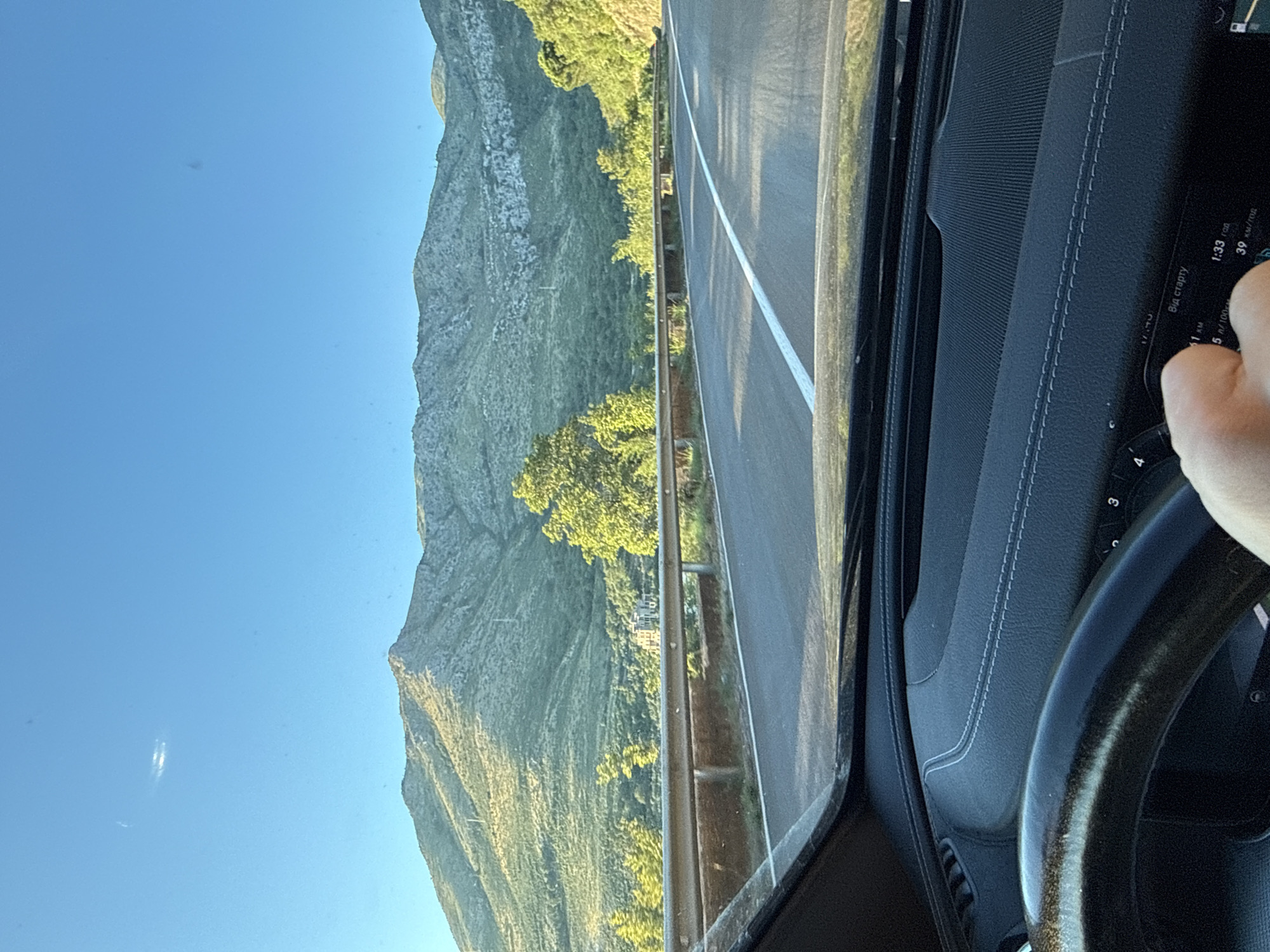 The image captures a driver's perspective from within a car, traveling on a road that winds through a mountainous landscape. The road, paved with asphalt, features a white dividing line. Guardrails made of metal line the side of the road. Lush green vegetation covers the slopes of the mountains, which are bathed in the warm, golden light of what appears to be late afternoon or early morning. A clear blue sky with a faint, elongated cloud is visible above. The interior of the car, including the steering wheel and dashboard, is partially visible in the foreground. A hand is holding onto the steering wheel. A digital display on the dashboard shows information including "Від старту" (From start), "1:33 год" (1:33 hours), and "39 км/год" (39 km/h). The scene is likely in Stupovi, Montenegro, given the characteristic mountainous terrain and vegetation.