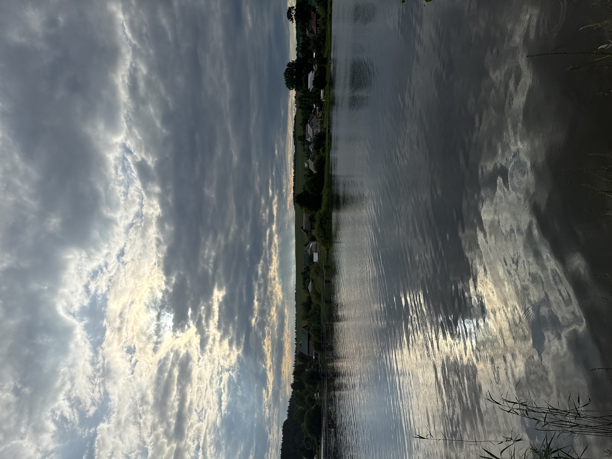 This is a landscape photograph taken in Luzhkovo, Russia, depicting a serene natural scene. The image is dominated by a large body of water, likely a river or lake, which occupies the foreground and mid-ground. The water's surface is rippled, reflecting the sky and the surrounding environment.
In the background, across the water, is a small village or settlement. Several houses with various roof colors, including green and brown, are visible, nestled amongst dense green trees. The vegetation appears lush, with rolling hills forming the backdrop to the settlement. A line of trees runs along the edge of the water in front of the village.
The sky above is dramatic, filled with a mix of dark, heavy clouds and brighter patches where sunlight is breaking through. The interplay of light and shadow creates a dynamic atmosphere. The overall impression is one of a peaceful, rural setting under a dynamic sky. There are no people or animals visible in the image.