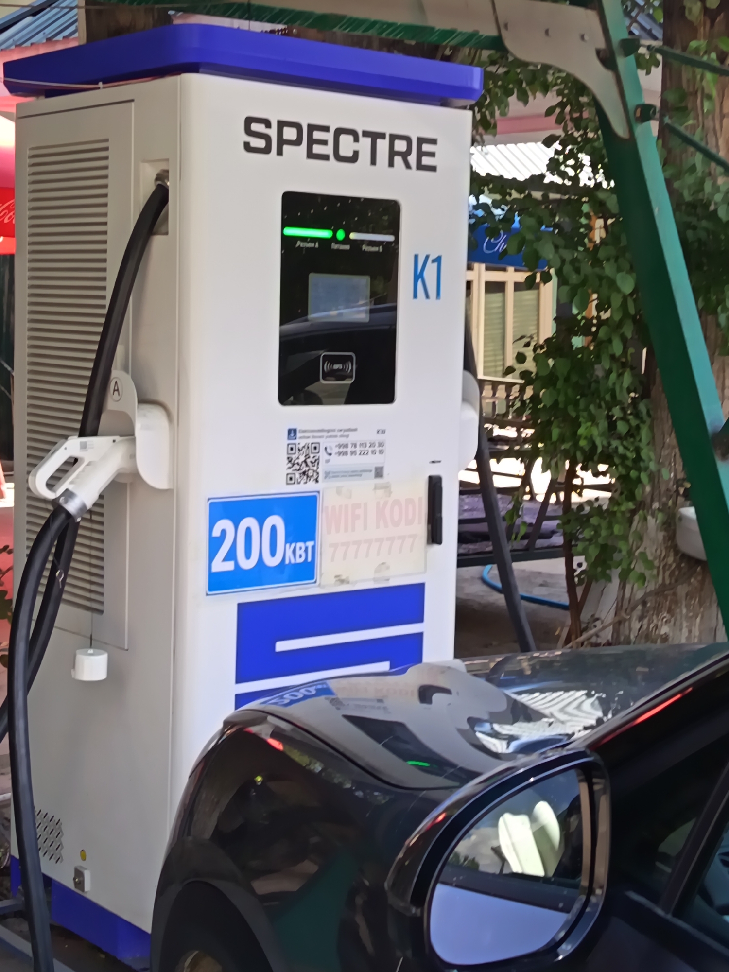 In the city of Rizaq, Uzbekistan, an electric vehicle charging station manufactured by "SPECTRE" and labeled "K1" is situated outdoors. The station, primarily white with blue accents, features a charging cable and connector labeled "A." A dark-colored car is parked next to the station, with its side mirror reflecting a portion of the charging station. The background consists of greenery, trees, and buildings, indicating an urban or semi-urban setting. Signs on the charging station display "200 KBT" and "WIFI KODI 7777777", along with QR codes and phone numbers. The scene appears to be captured during daylight.