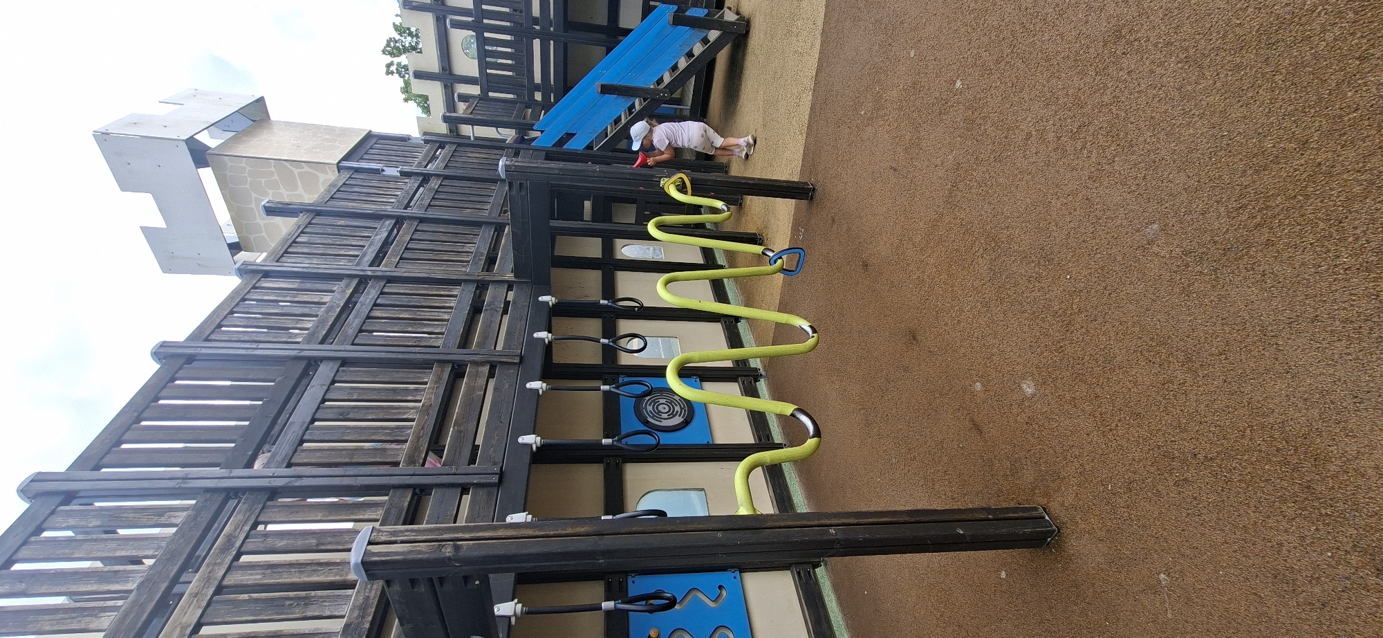 This image depicts a children's playground structure, likely designed to resemble a castle, situated in Lahti, Finland. The structure is primarily constructed of dark wood, with wooden planks forming railings and vertical supports. A multi-level platform is visible, with a blue slide extending from an upper level to the ground. Attached to the underside of the upper level are several black hanging rings. A wavy, bright yellow tube-like structure, also with hanging rings, extends across the playground. The ground surface of the playground is a textured, reddish-brown material, possibly rubber mulch or a similar safety surface. A young child, wearing a light-colored hat and clothing, is standing near the blue slide, appearing to interact with a red object. The sky is overcast. The overall environment suggests an outdoor recreational area designed for children's play.