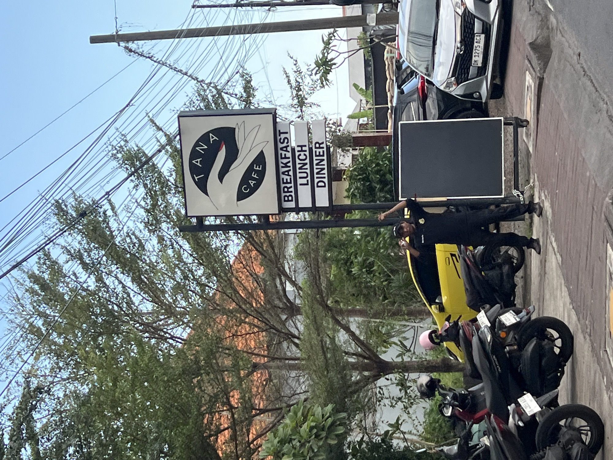 The image shows the exterior of Tana Cafe in North Kuta, Indonesia. A black-clad individual is standing beside a sign that advertises "Breakfast, Lunch, Dinner." Several parked motorbikes are on the sidewalk. A silver-colored car with the license plate "DK 1275 ACA" is parked nearby. Utility lines are visible overhead, against a clear sky. Trees and vegetation surround the cafe sign.