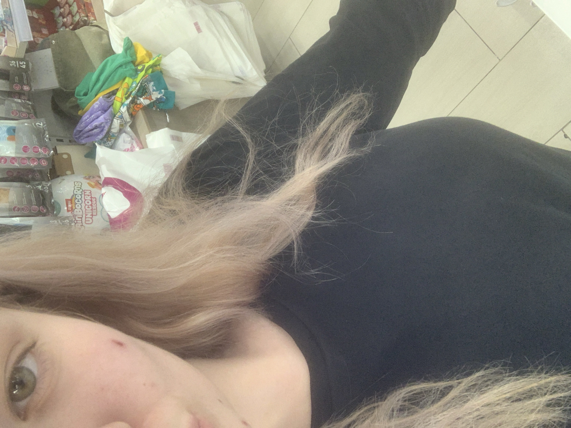 This close-up, angled media file features a person with long, blonde hair, partially obscuring their face on the left side of the frame. One eye, with a light iris (appearing green or hazel), is visible, along with part of a cheek that shows a faint reddish mark. The person is wearing a dark-colored garment, likely a black long-sleeved top or sweatshirt, which fills a significant portion of the image.
The background depicts an indoor retail environment, resembling an aisle in a supermarket or department store. The floor is covered with light-colored tiles. On the left, shelving units display various packaged products, with one label clearly showing the numbers "36 45". In the mid-ground, a stack of colorful fabric items or clothing (including green, yellow, purple, and patterned pieces) is visible, alongside several white shopping bags. A prominent white and pink package, labeled "Rain BocoRns UNICORN RESCUE", is also in the background.
The image captures a casual moment, possibly a selfie taken while shopping. No specific activity beyond the act of photography is discernible. The lighting suggests typical daytime or evening store hours. The location cues, such as shelves of products and shopping bags, confirm a commercial setting within Saint Petersburg, Russia.