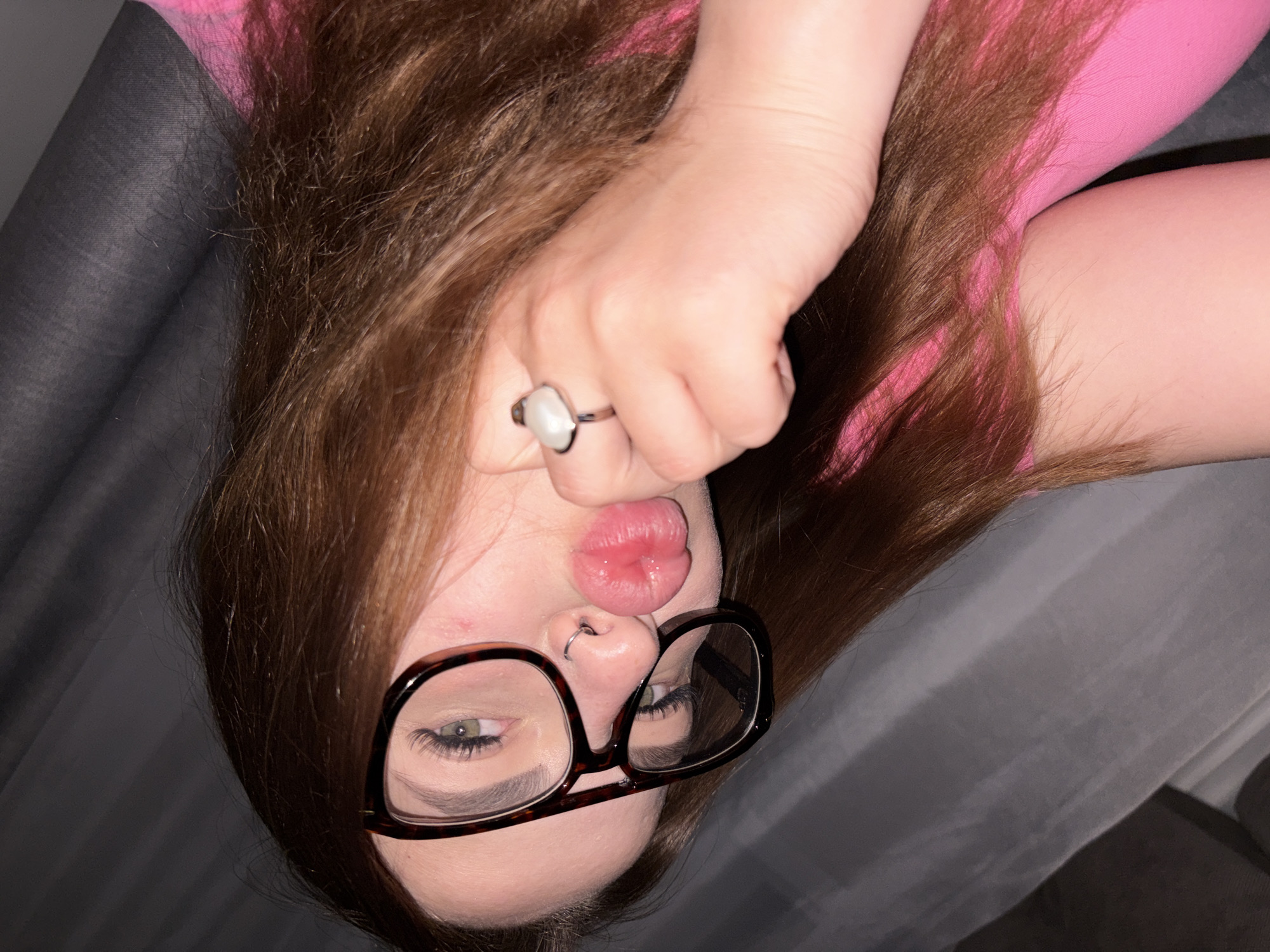 A woman with long brown hair and wearing black-rimmed glasses is shown upside down. She has a nose ring and is wearing a ring on her finger. She is puckering her lips. She is wearing a pink top. The background appears to be gray fabric. It is not possible to determine the location is Krasnodar, Russia, from the given image.
