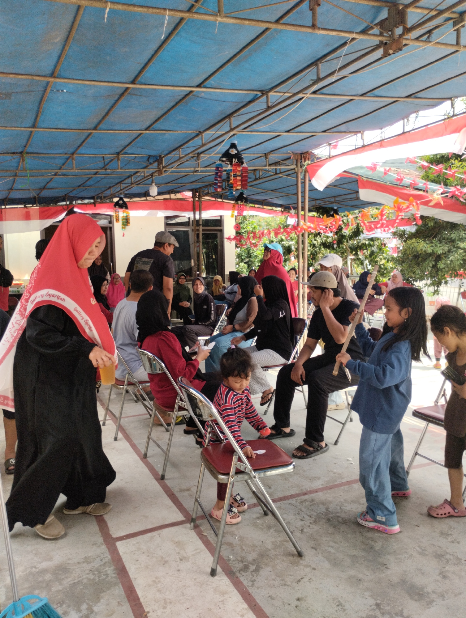 The image shows an outdoor gathering under a blue tarp supported by a metal frame. Red and white Indonesian flags are draped along the structure, along with colorful festive decorations. A group of people, including adults and children, are seated and standing. In the foreground, a woman wearing a red hijab and a black dress holds a clear cup. To her right, a young girl in a blue jacket is holding a wooden stick. Other individuals are seated on metal chairs, some engaged in conversation or looking at their phones. The ground is a concrete surface with red lines marking sections. The overall scene suggests a community event or celebration.