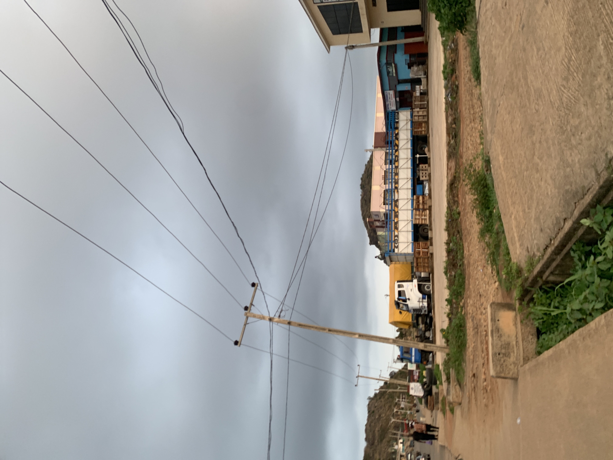 The image captures a street scene in Gura Top, Nigeria, under a heavy, overcast sky. The street is lined with buildings on one side and a concrete barrier with sparse vegetation on the other. A series of utility poles with power lines stretch across the frame. Several trucks, one white with a yellow cover, are parked along the street. In the distance, a hill is visible. Some pedestrians are walking further down the road.