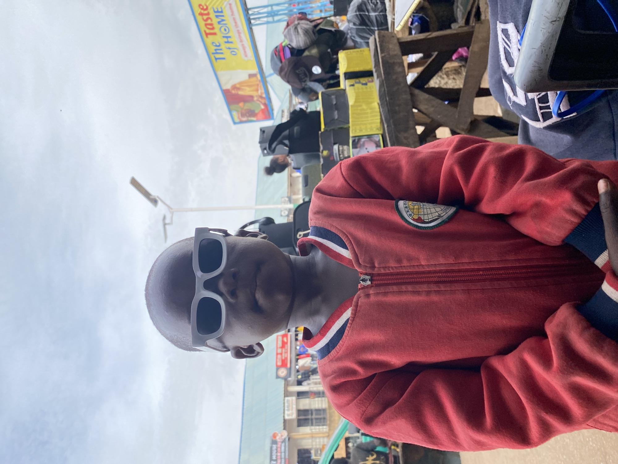 The image depicts a person wearing sunglasses and a red jacket with a patch on the left sleeve. The individual is positioned in an outdoor environment, likely a market or public space, given the presence of various stalls, signs, and other individuals in the background. The location appears to be Bukuru, Nigeria, based on the "The Taste of HOME" sign, which is a local business. The overall scene suggests a casual, everyday moment captured in a bustling urban setting.