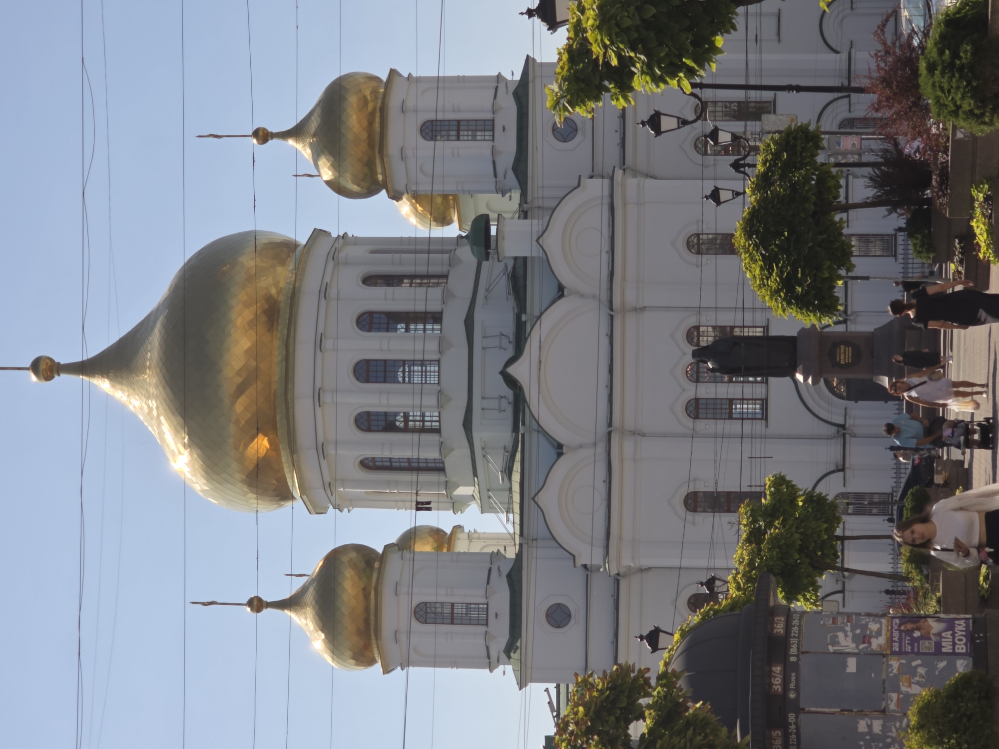The image shows a large, white Orthodox church with prominent golden domes, characteristic of Rostov-on-Don, Russia. The church has multiple domes, with the central one being the largest and most ornate, topped with a cross. Smaller domes are visible on flanking towers. The building's architecture features arched windows and classical detailing. In the foreground, there is a pedestrian walkway with several people walking. A woman in a white top and dark skirt is looking at her phone, and another woman in a black dress is walking away from the camera. A man in a blue shirt is pushing a stroller, and a woman in a white dress is walking beside him. To the left of the walkway, there is a black kiosk with advertisements and posters. Several trees and decorative bushes are planted along the walkway, and lampposts are visible. The sky is clear and blue, suggesting a sunny day. The overall scene captures a public space in a city, with religious architecture and everyday life coexisting.