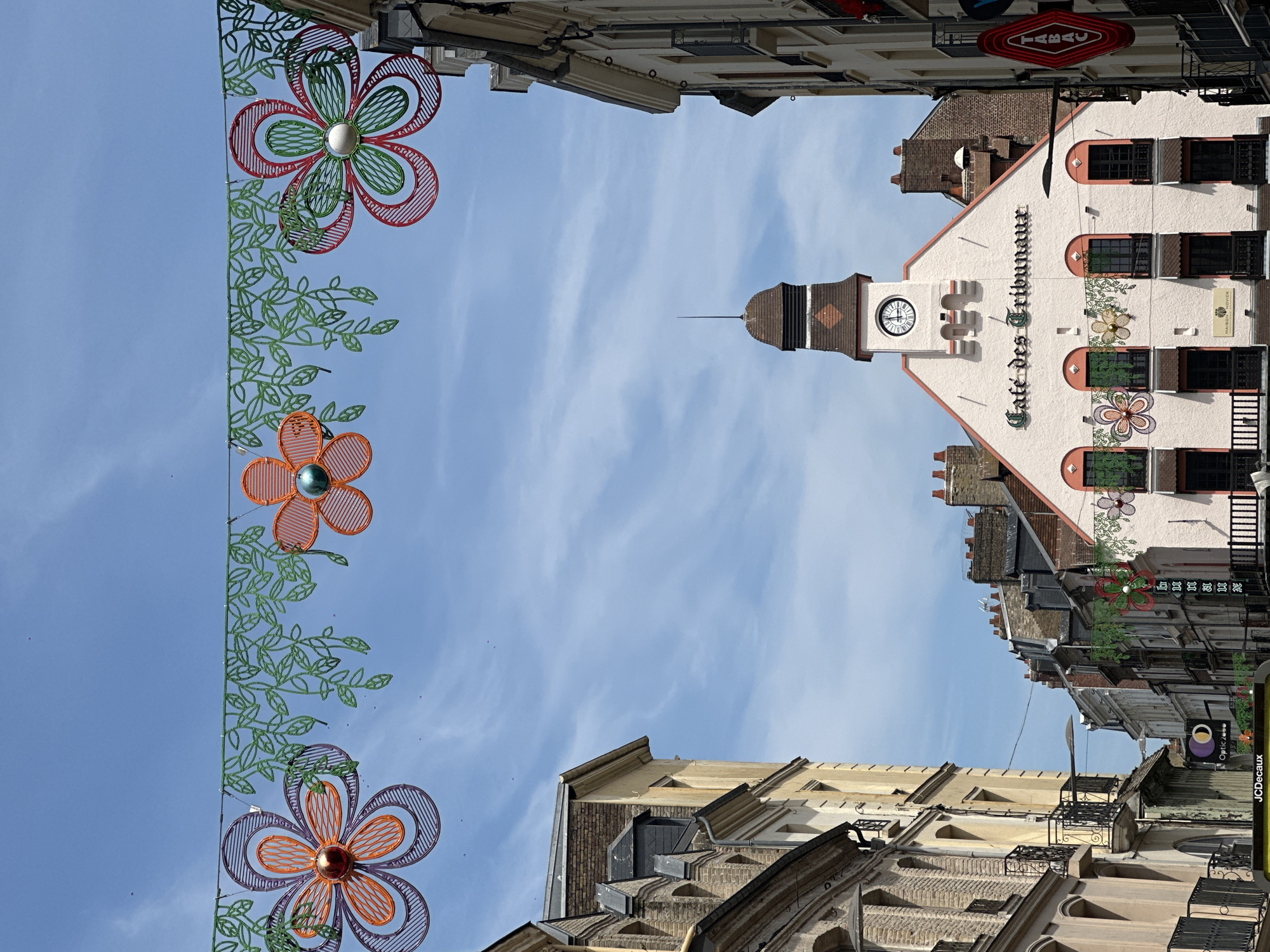 This is a daytime, eye-level, medium shot of a street scene in Dieppe, France, featuring the Café des Tribunaux building. The image is dominated by the sky, which is a bright blue with wispy white clouds. Hanging across the top of the frame are decorative garlands of green leaves and three large, colorful wire flowers, one purple and orange on the left, one orange in the center, and one red and green on the right.
The Café des Tribunaux building is a prominent, pale pink structure with a clock tower. The clock face shows the time as approximately 12:30. The building has several windows with dark frames and awnings, and ornate trim. Below the clock, there are decorative floral elements. The building's name, "Café des Tribunaux", is written in elegant script above the windows.
On either side of the Café des Tribunaux are buildings typical of a European city street, with brick and stone facades, arched windows, and balconies. Some of the buildings appear older, with weathered brickwork. A red neon sign reading "TABAC" is visible on the right side of the frame. In the lower left corner, a portion of a yellow sign with "JCDecaux" is visible, indicating a public advertising service. There are no people clearly visible in the image. The overall atmosphere is bright and sunny, suggesting a pleasant day in the city.