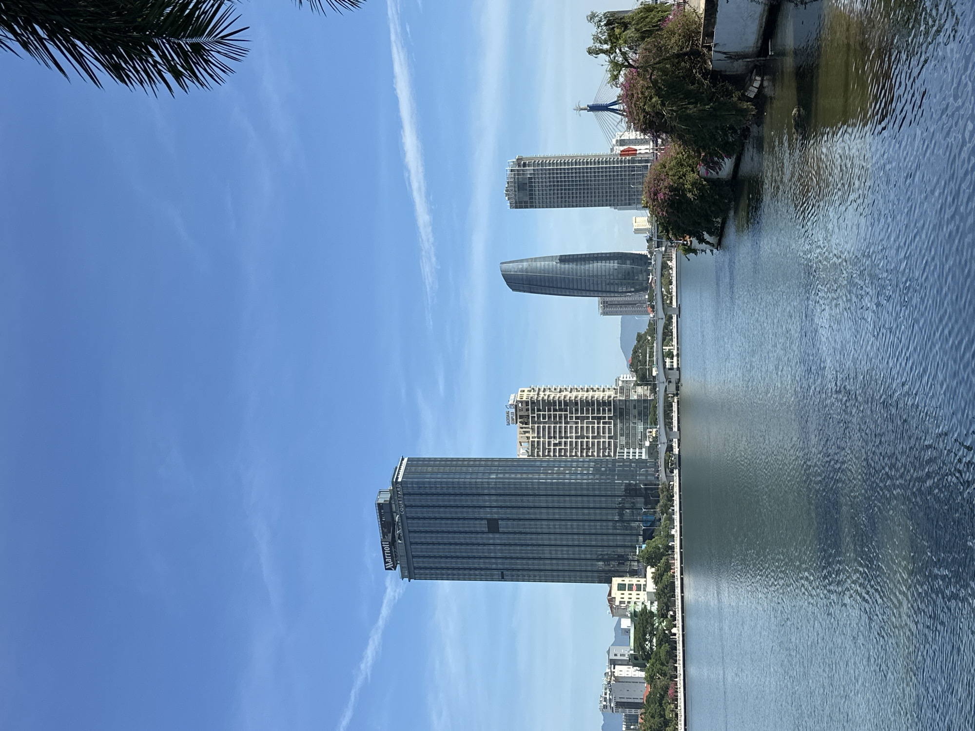 A low-angle, landscape-oriented shot captures a waterfront cityscape under a bright, clear blue sky with wispy white clouds. A palm frond is visible in the upper left corner. The scene is dominated by a large body of water on the right, reflecting the sky and the buildings on the opposite bank.
Several modern skyscrapers line the waterfront. In the foreground on the left, a tall, rectangular building with a dark, glass facade is prominent. A sign on its upper section reads "COURTYARD MARRIOTT." Further back and to the right, another sleek, curved skyscraper made of dark glass stands beside a more conventionally designed, grid-like tall building. Other buildings of varying heights and architectural styles are visible further down the coastline, suggesting a developed urban area.
The water exhibits gentle ripples, indicating calm conditions. On the bank, lush green trees, some adorned with purple flowers, provide a natural contrast to the urban architecture. A bridge structure, featuring cables and towers, is visible in the distance, adding to the sense of a thriving city. The overall impression is one of a sunny day in a bustling coastal city, likely in the afternoon due to the quality of light. The image conveys a sense of urban development and natural beauty coexisting.