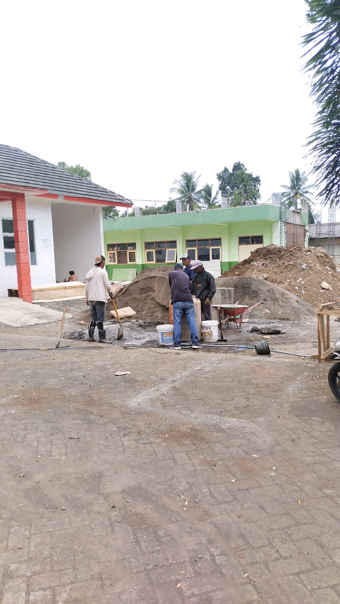 In Klungkung, Indonesia, a construction site is captured under an overcast sky. A building with a white and red facade and a tiled roof is situated on the left, adjacent to a green building with multiple windows. In the foreground, several workers are engaged in construction activities. One worker, wearing a light-colored jacket, a cap, and dark boots, holds a shovel next to a pile of sand. Three other workers are gathered around a cement mixer, actively working with materials. Buckets, some labeled "JOTUN" and "WIFEX," are present, along with a wheelbarrow and various construction tools scattered around the site. Piles of sand and dirt dominate the central area, indicating ongoing excavation and building. The ground is a mix of dirt and paving stones. In the background, palm trees and other foliage suggest a tropical environment.